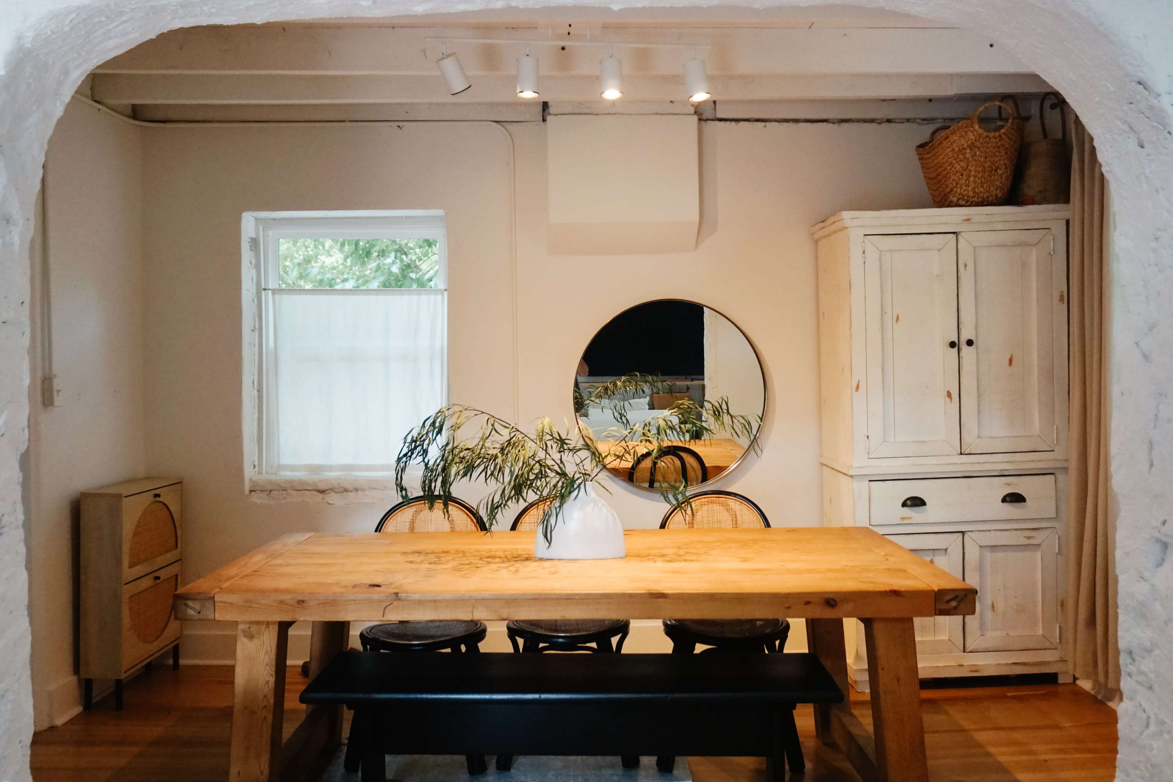 A wooden dining table with chairs is centered in a room featuring a round mirror and a white cabinet against the wall.