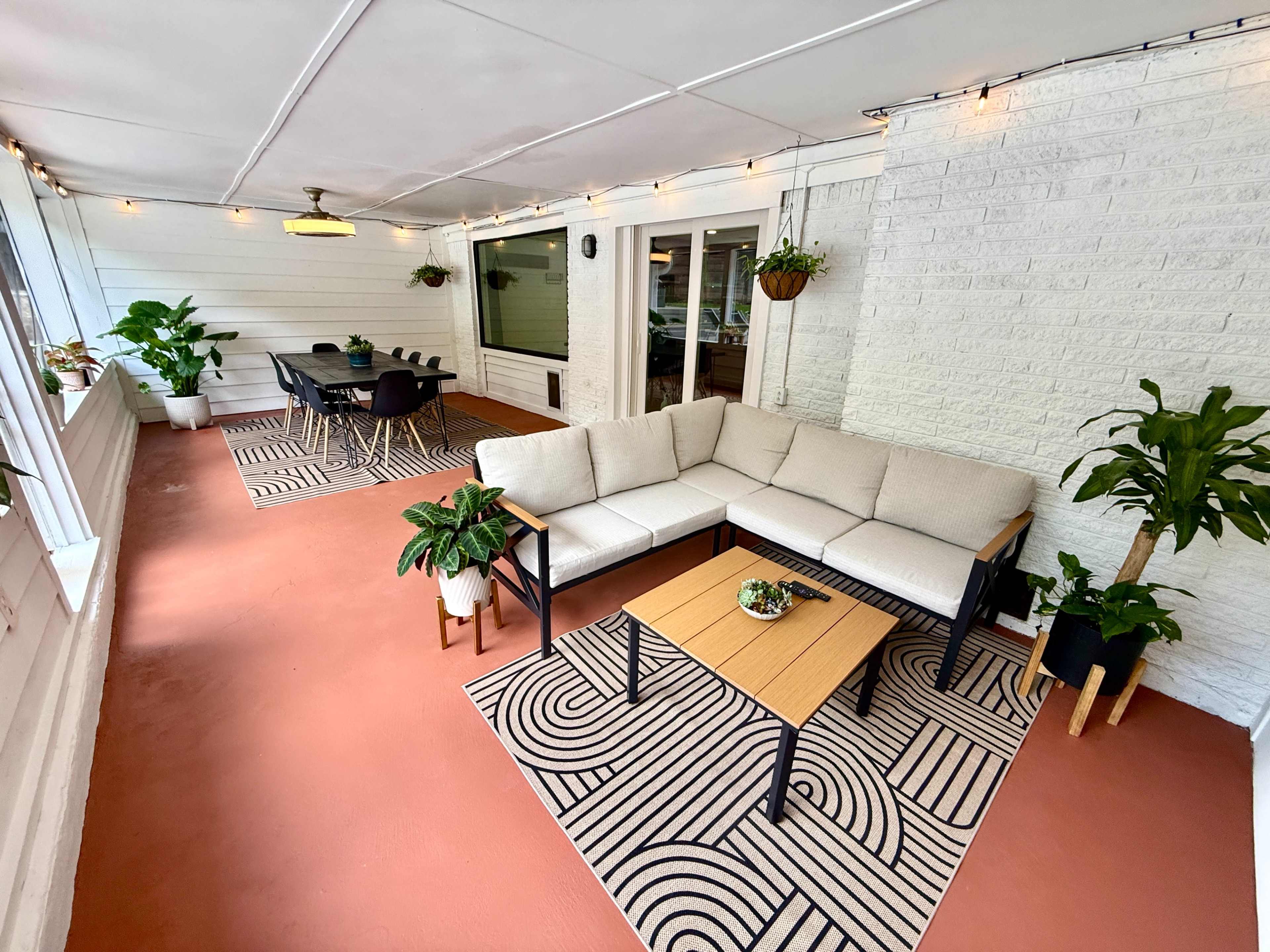 A bright sunroom features a sectional sofa, a coffee table, and a dining area adorned with potted plants and overhead string lights.