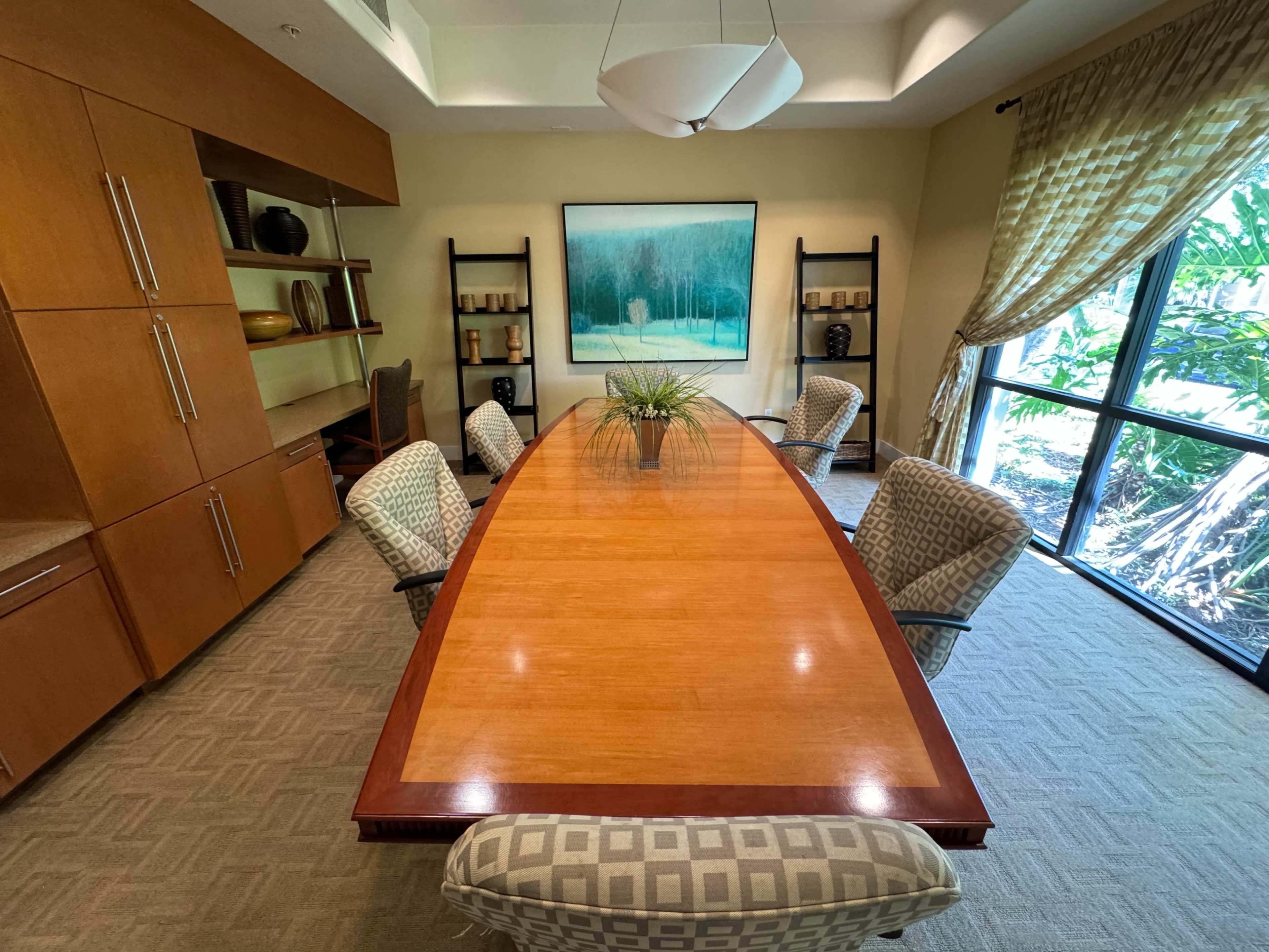 A modern conference room features a long wooden table surrounded by upholstered chairs, with a large artwork on the wall and windows showing greenery outside.