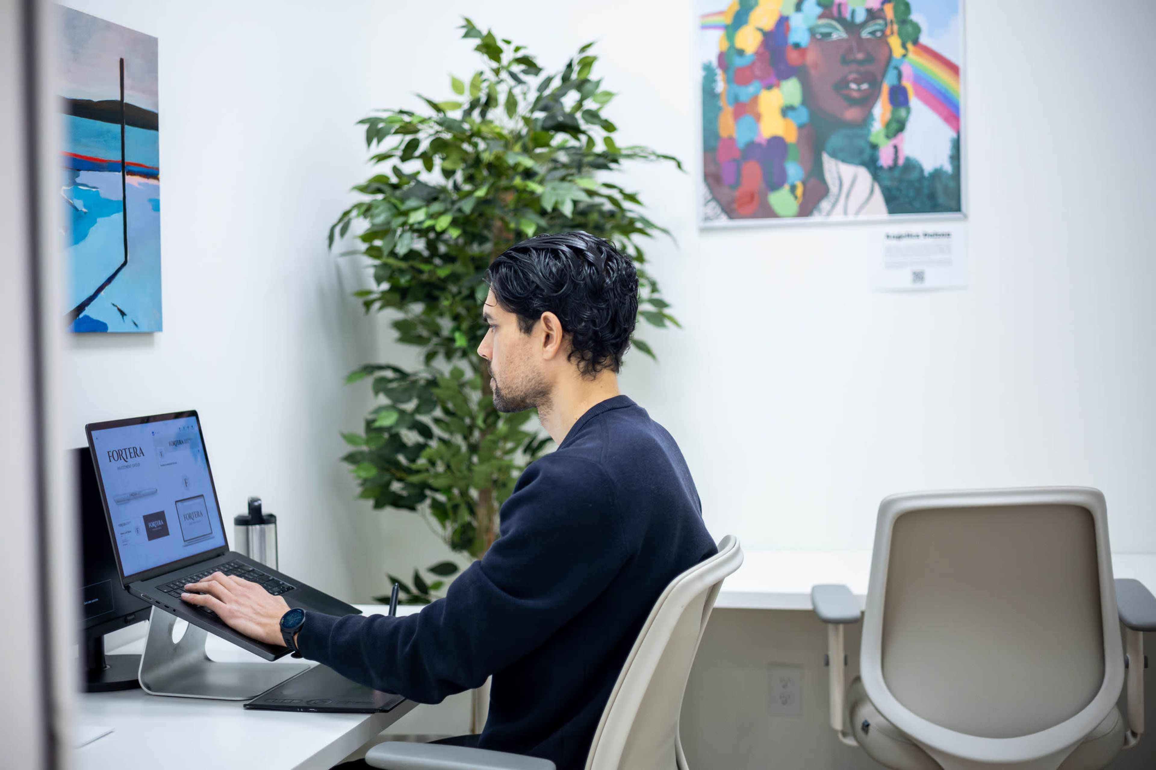 A man works on a laptop at a desk in a well-lit room with a plant and colorful artwork on the walls.