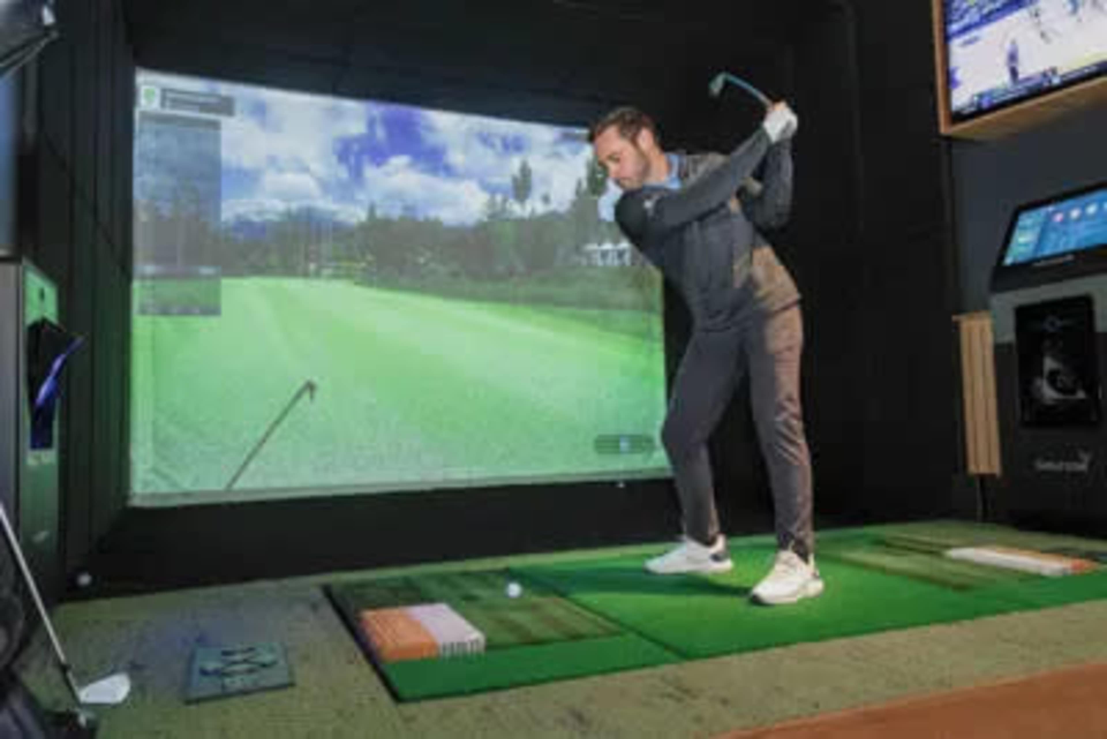 A person is using a golf simulator, swinging a club in front of a large screen displaying a golf course.