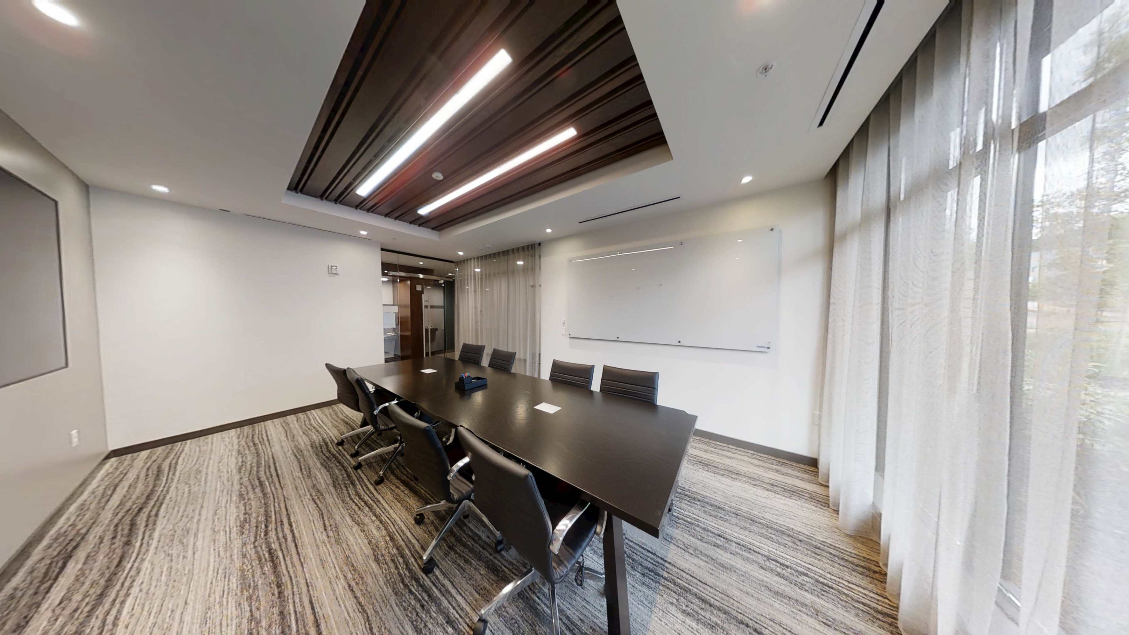 The image shows a modern conference room featuring a long dark table surrounded by several black chairs, with a large whiteboard and floor-to-ceiling windows.