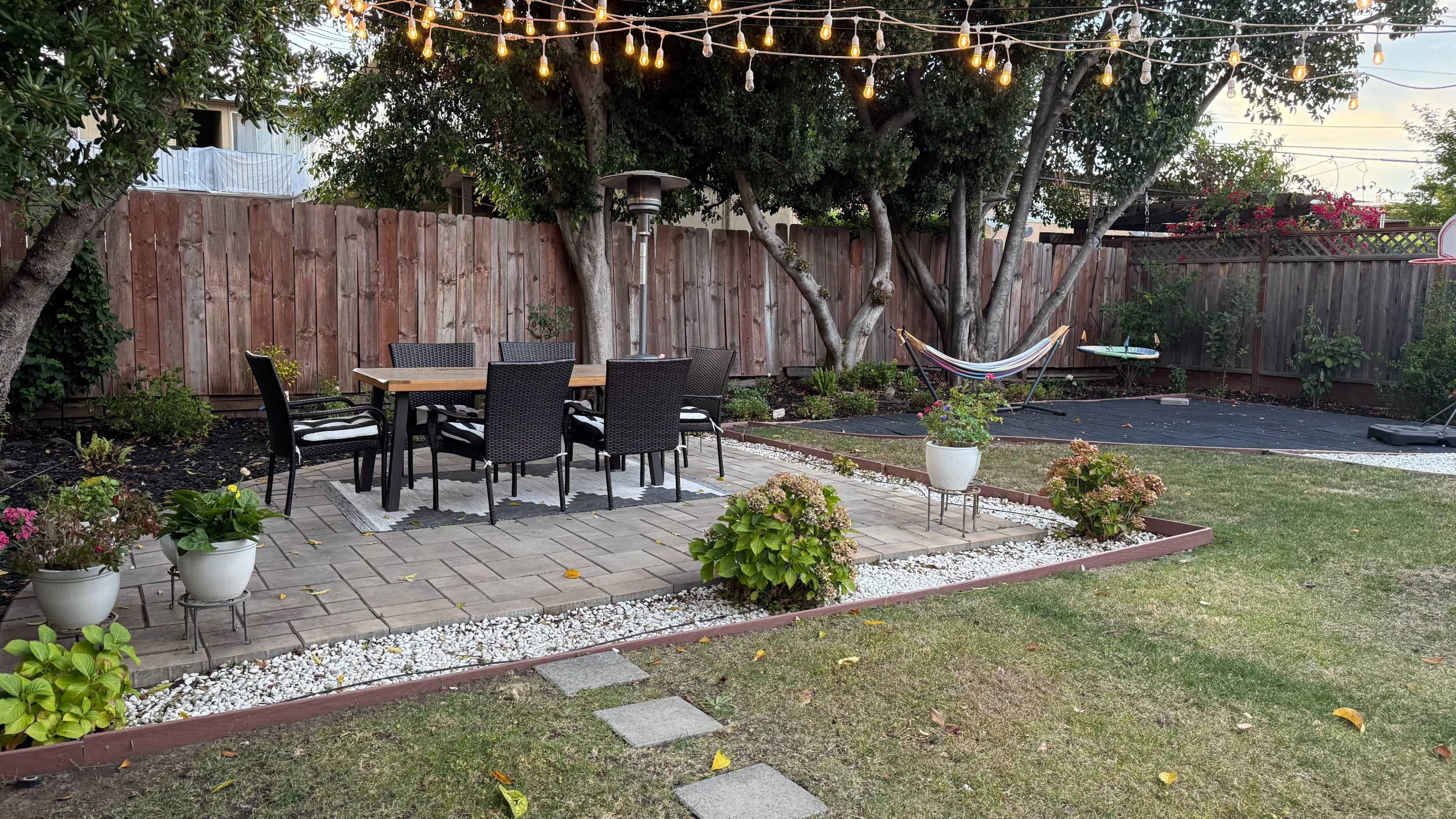 The image shows a landscaped backyard with a dining table and chairs, string lights overhead, a hammock, and flower pots along a stone pathway.