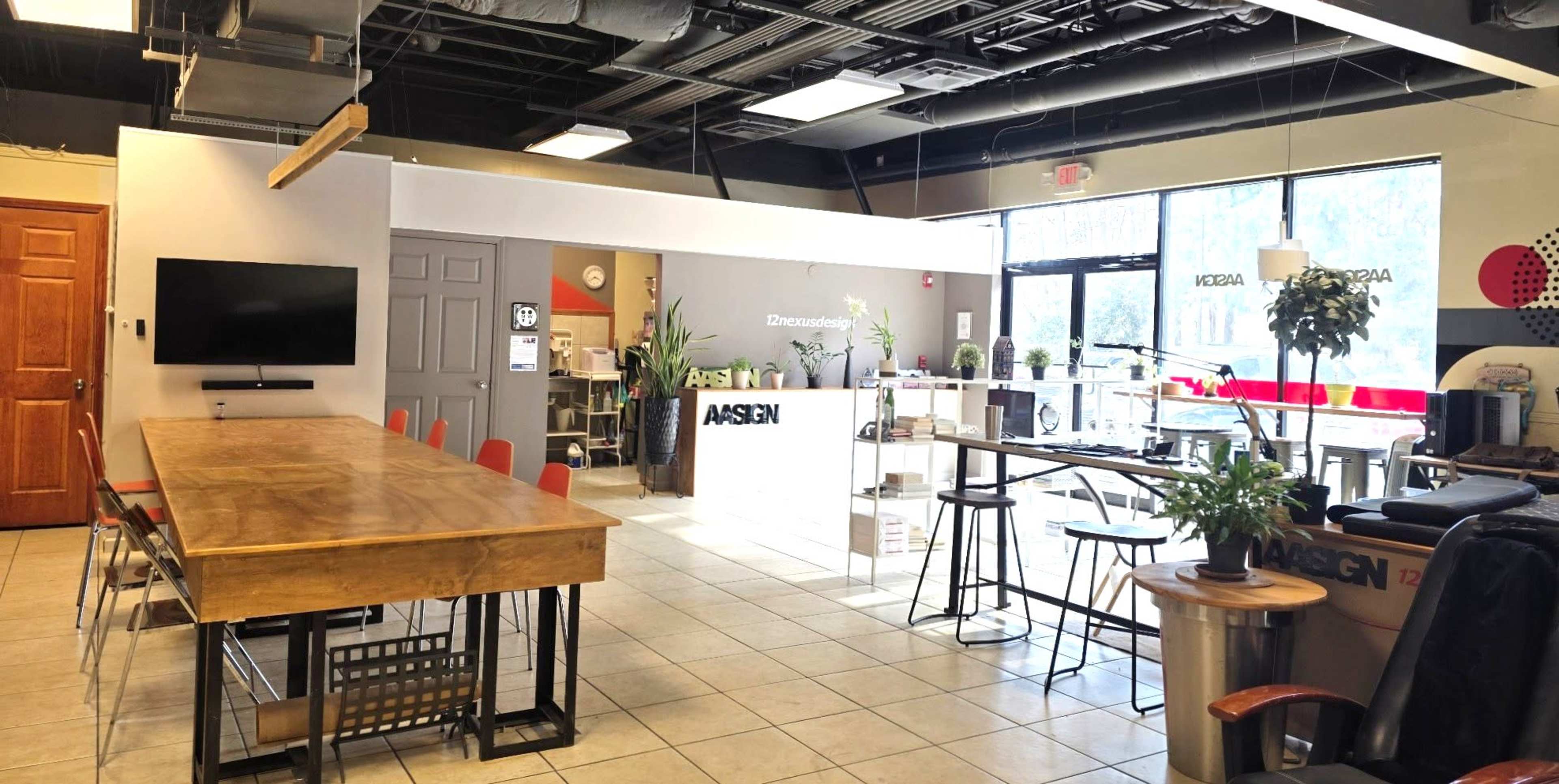 The image shows a modern office space with a long wooden table, various seating areas, and large windows allowing natural light to enter.