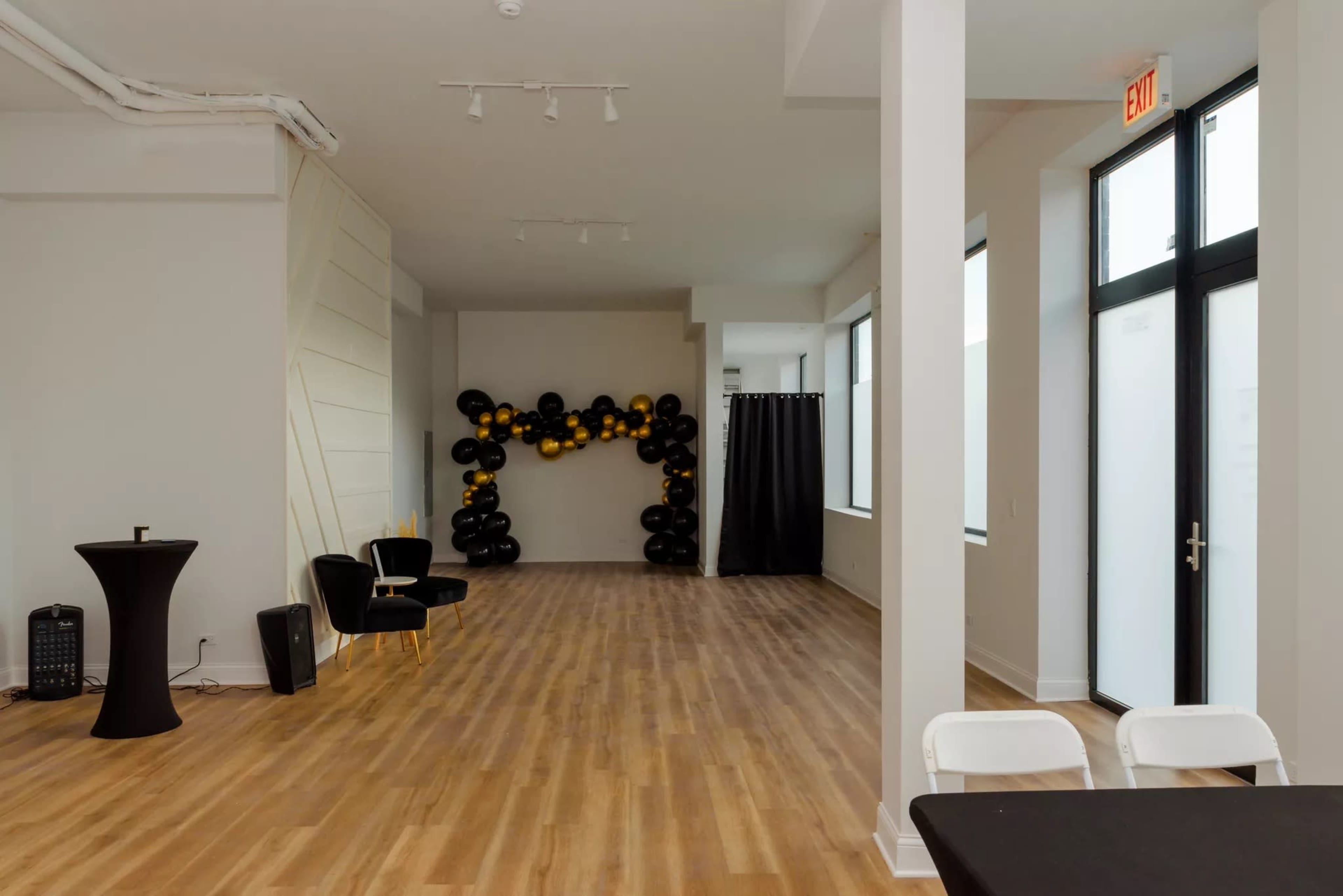 A spacious room with wooden flooring, featuring a black and gold balloon decoration on one wall, a black round table, two chairs, and a closed curtain in the background.
