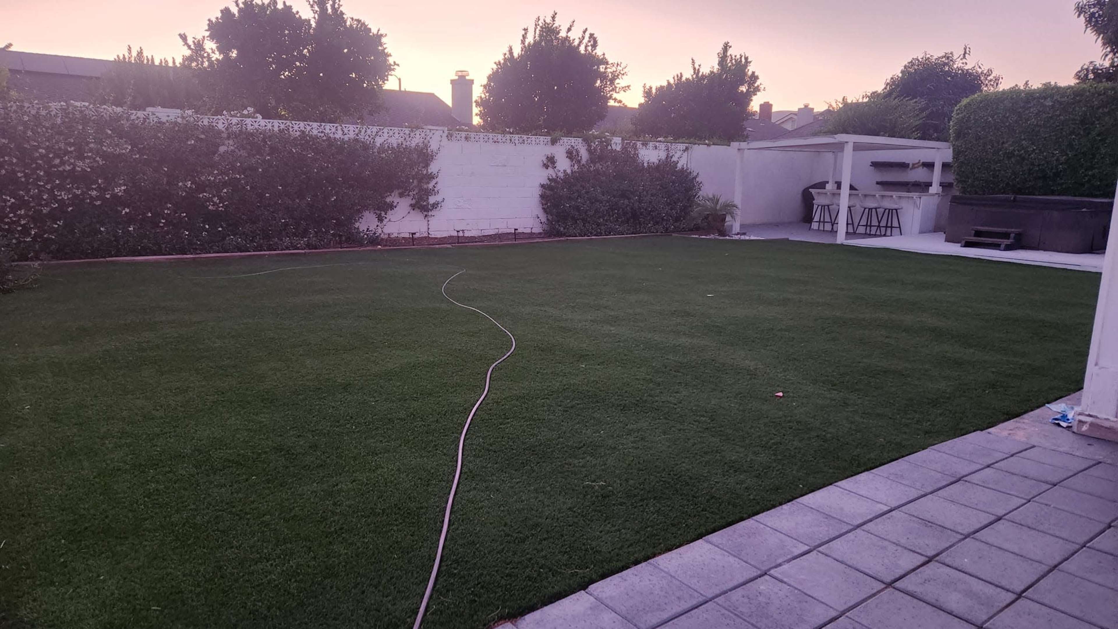 The image shows a large, well-maintained backyard with artificial turf, a paved area, and greenery along the perimeter, under a pinkish sky at dusk.