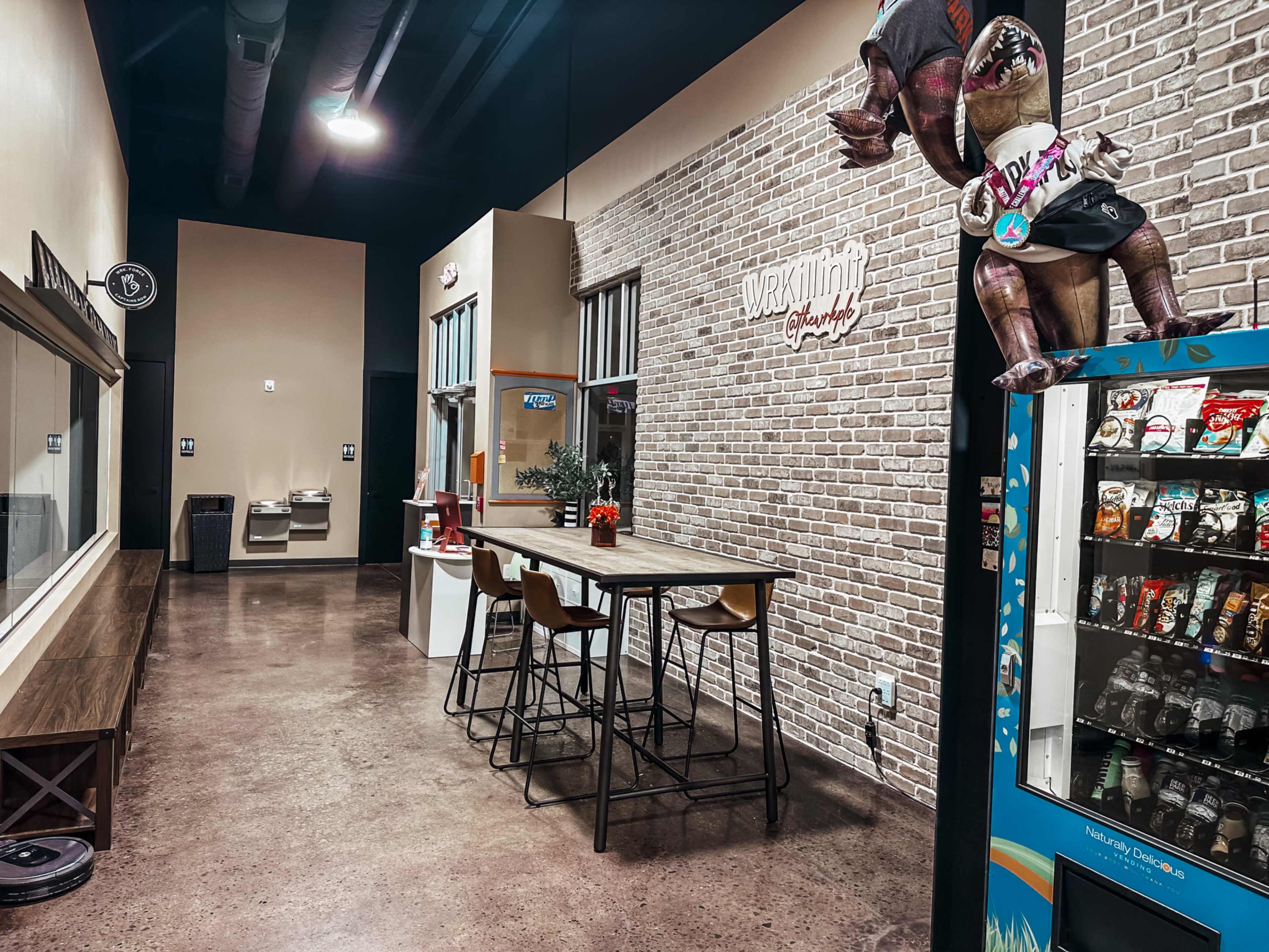 The image shows a modern interior space featuring a long table with high stools, a vending machine, and a decorative wall with a character figure.