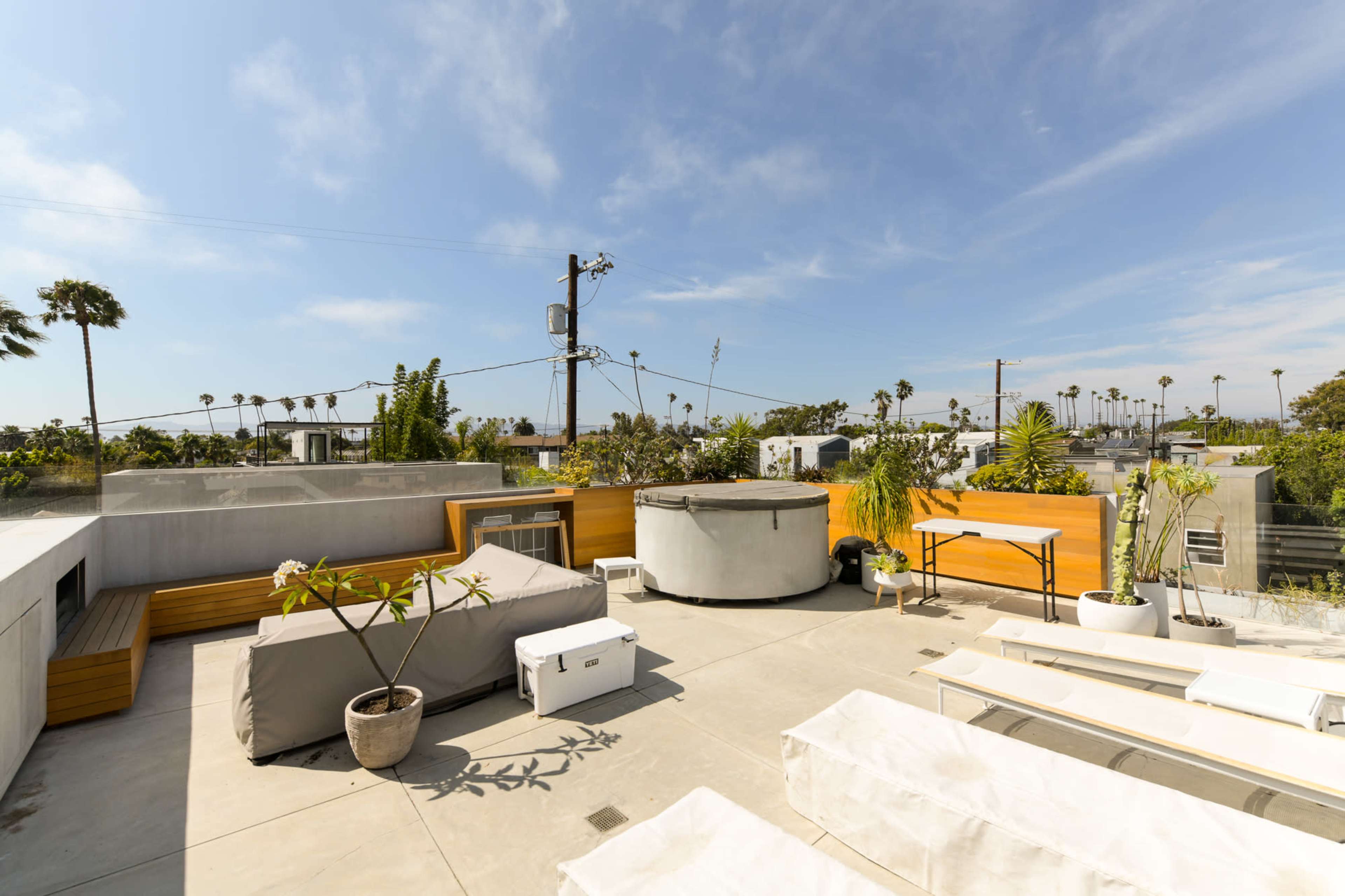 The rooftop terrace features wooden seating, a round hot tub, and several potted plants against a backdrop of palm trees and blue sky.