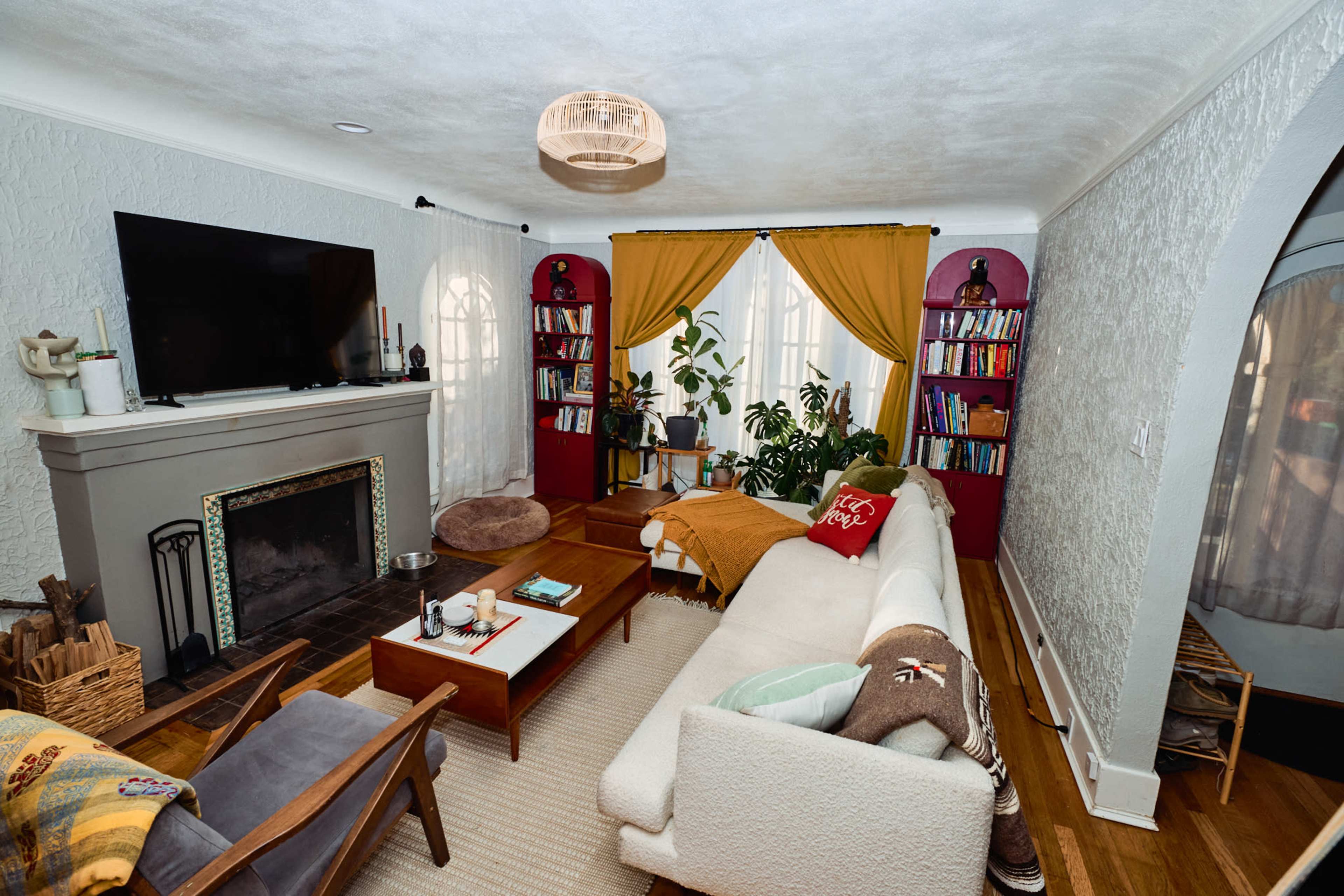 The living room features a large television mounted above a fireplace, a sectional sofa, bookshelves filled with books, and potted plants set against a backdrop of yellow and maroon curtains.