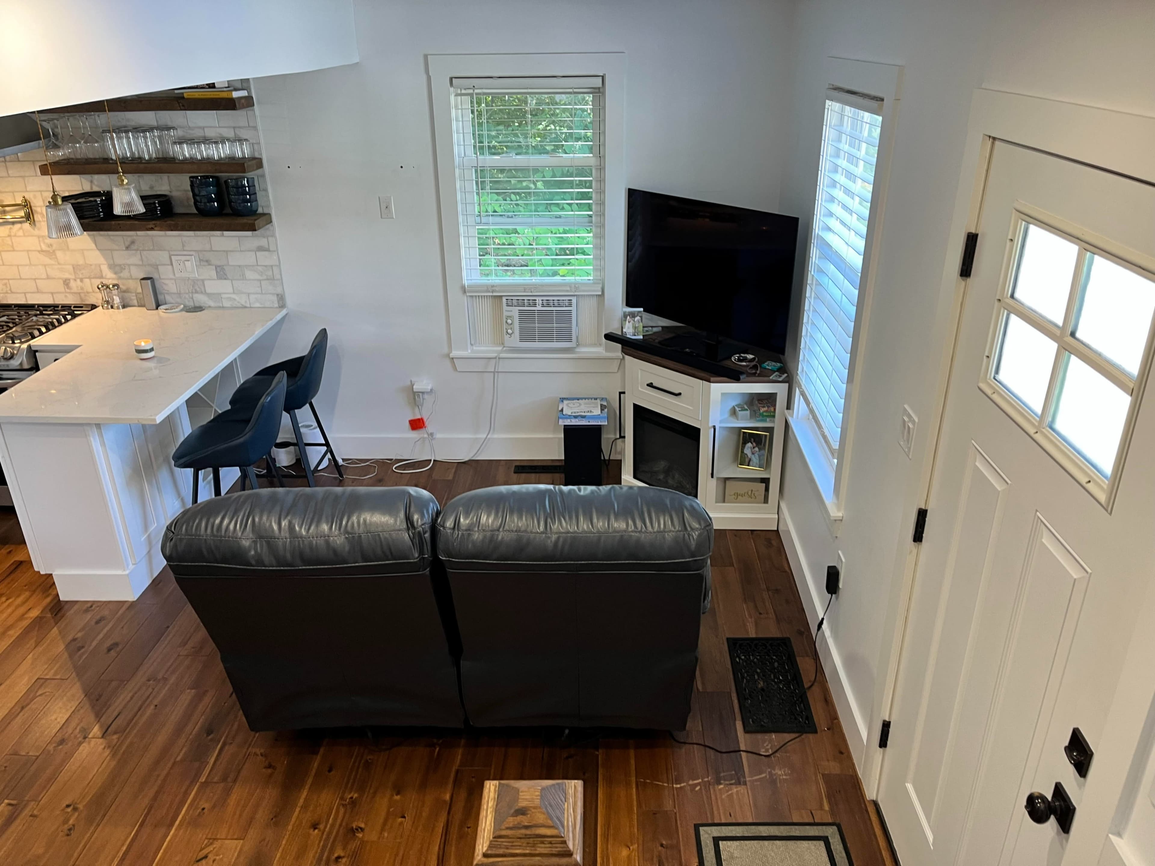 The image shows a cozy living space with a gray couch facing a television on a cabinet, an air conditioning unit by the window, and a kitchen area visible in the background.