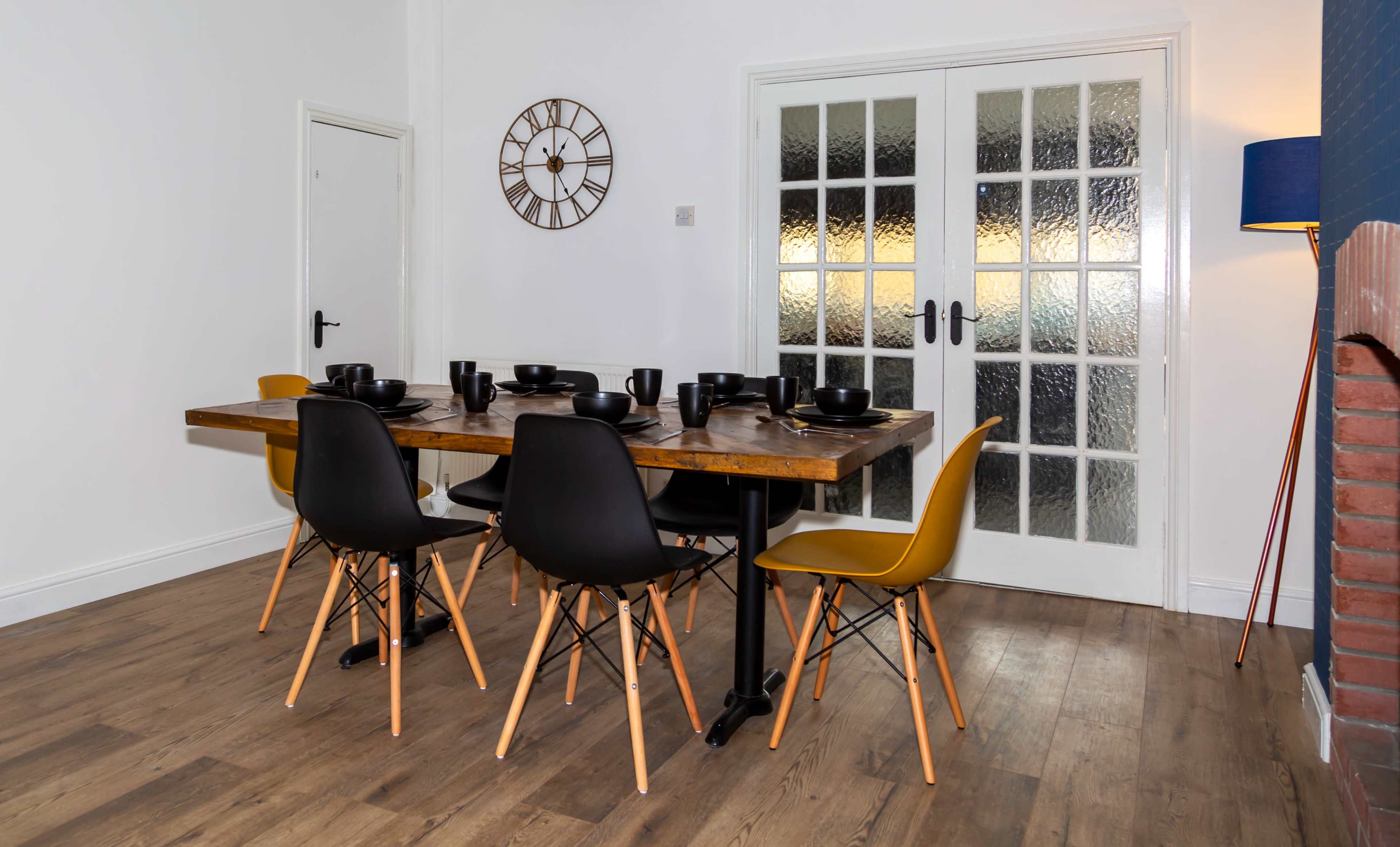 A rectangular wooden dining table is set with black bowls, surrounded by a mix of black and yellow chairs, in a room featuring a clock on the wall and glass-paneled doors.