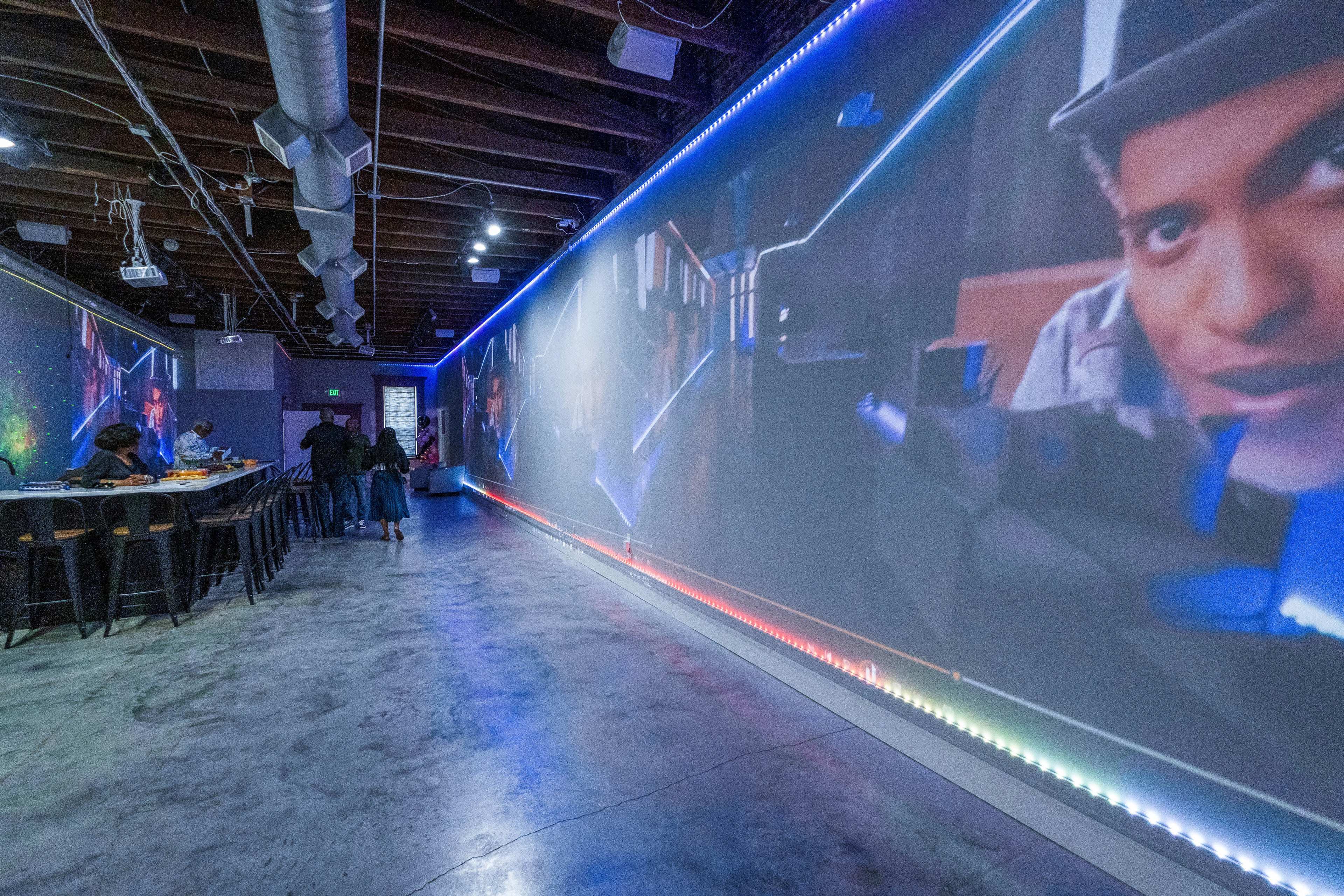 A spacious indoor area with a long wall displaying vibrant digital screens and several people seated at high tables.