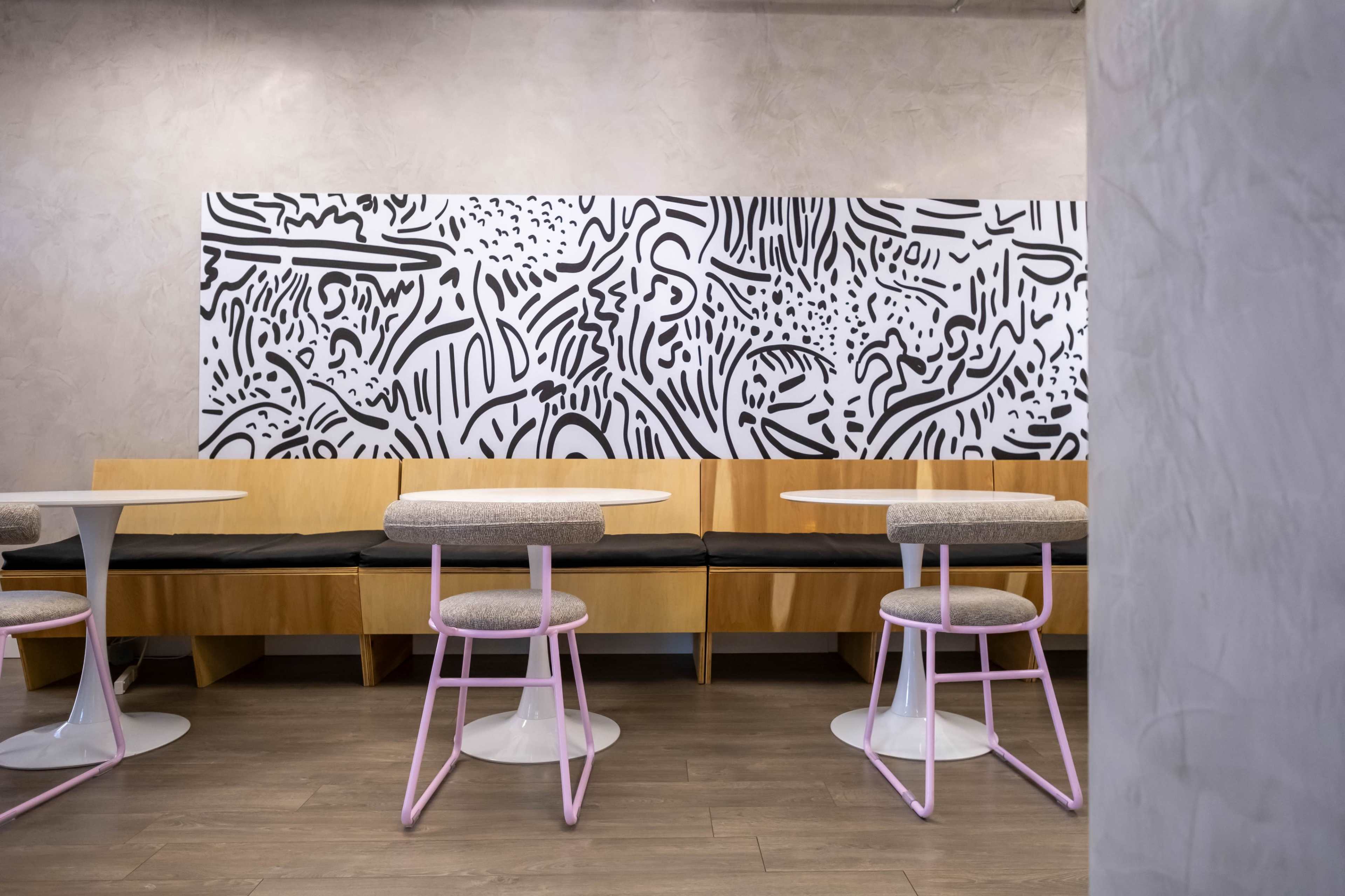 The image shows a modern cafe interior featuring wooden seating, round tables, and a large black and white abstract mural on the wall.
