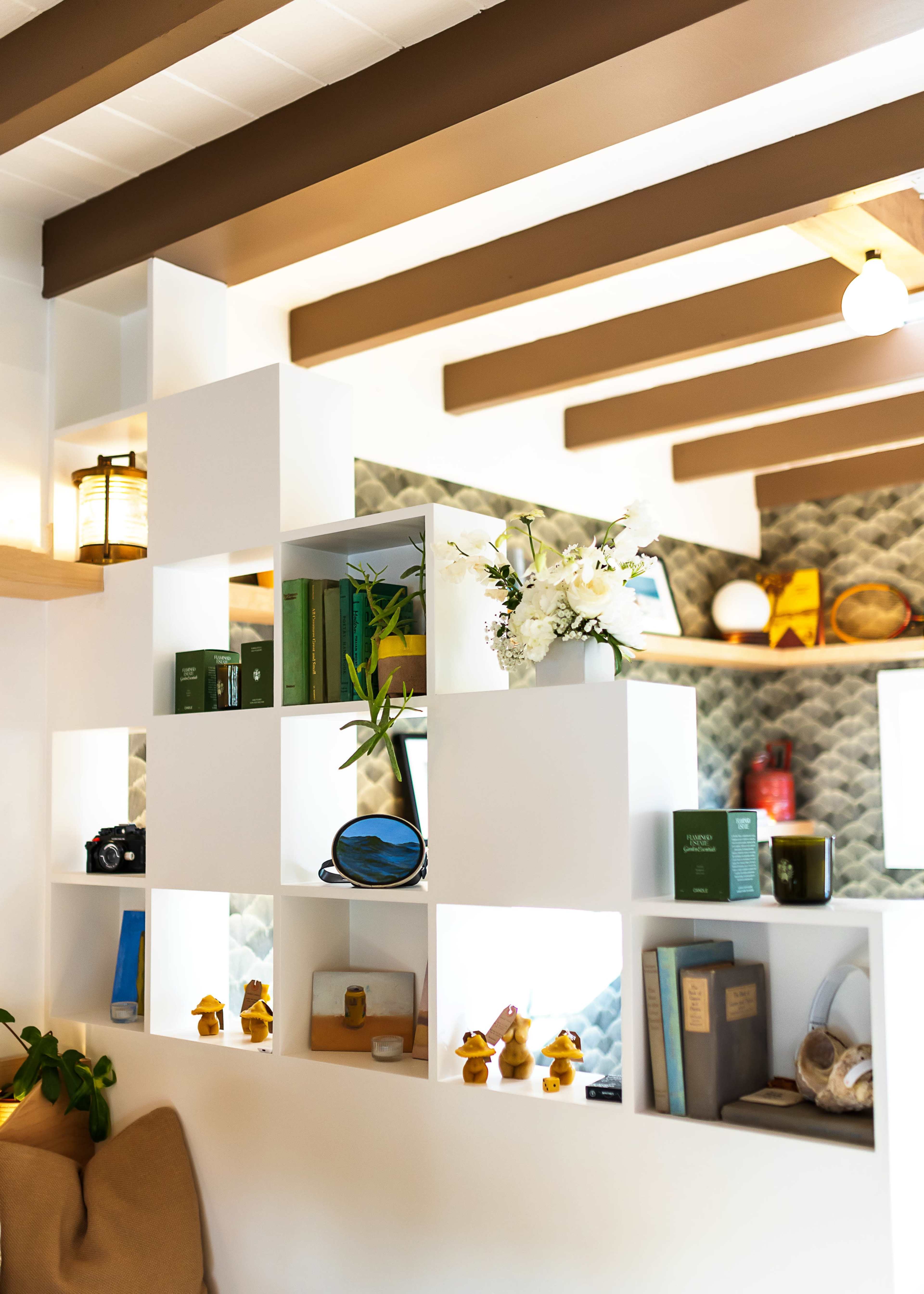 A white shelving unit with various decorative items, including books, a camera, and plants, set against a patterned wall and wooden beams.