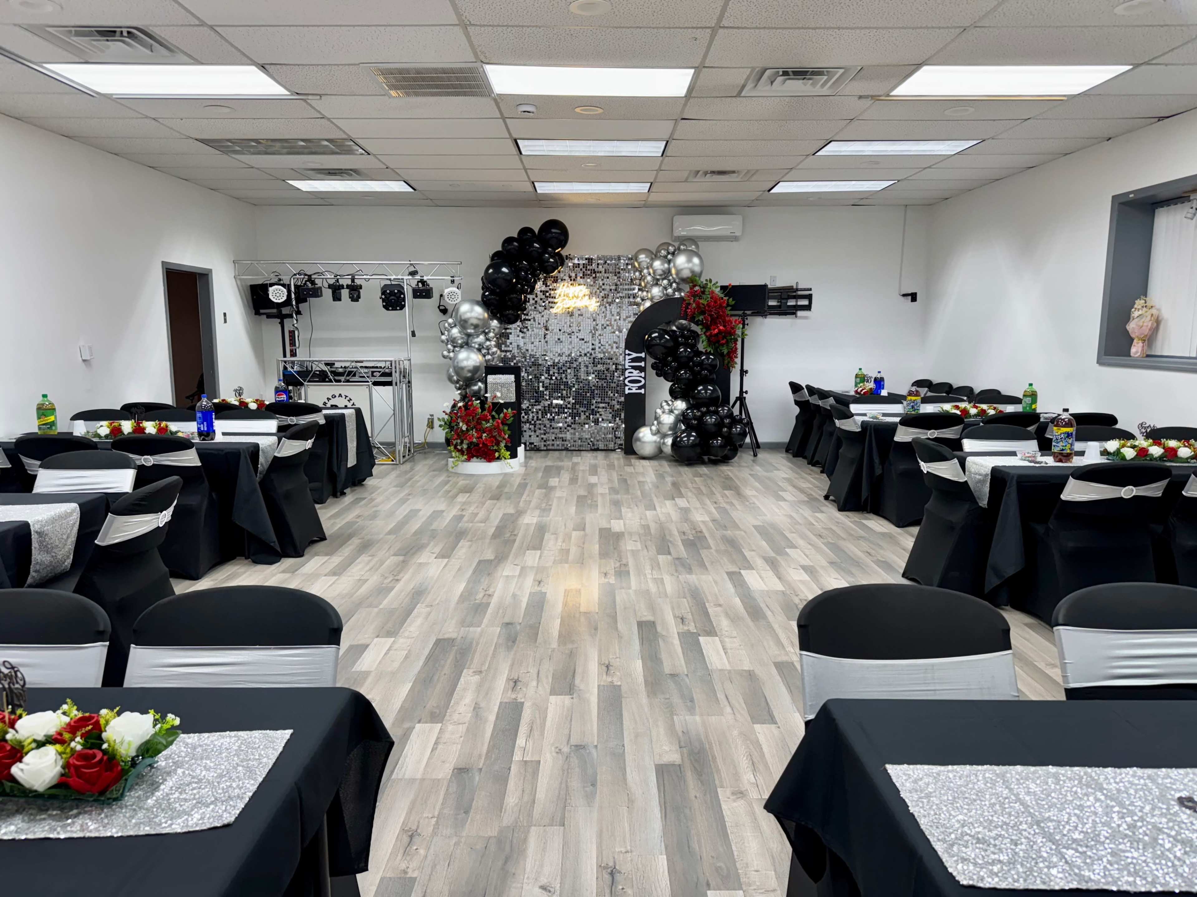 The image shows a decorated event space with tables set up, featuring black tablecloths, floral centerpieces, and a backdrop of balloons and streamers.