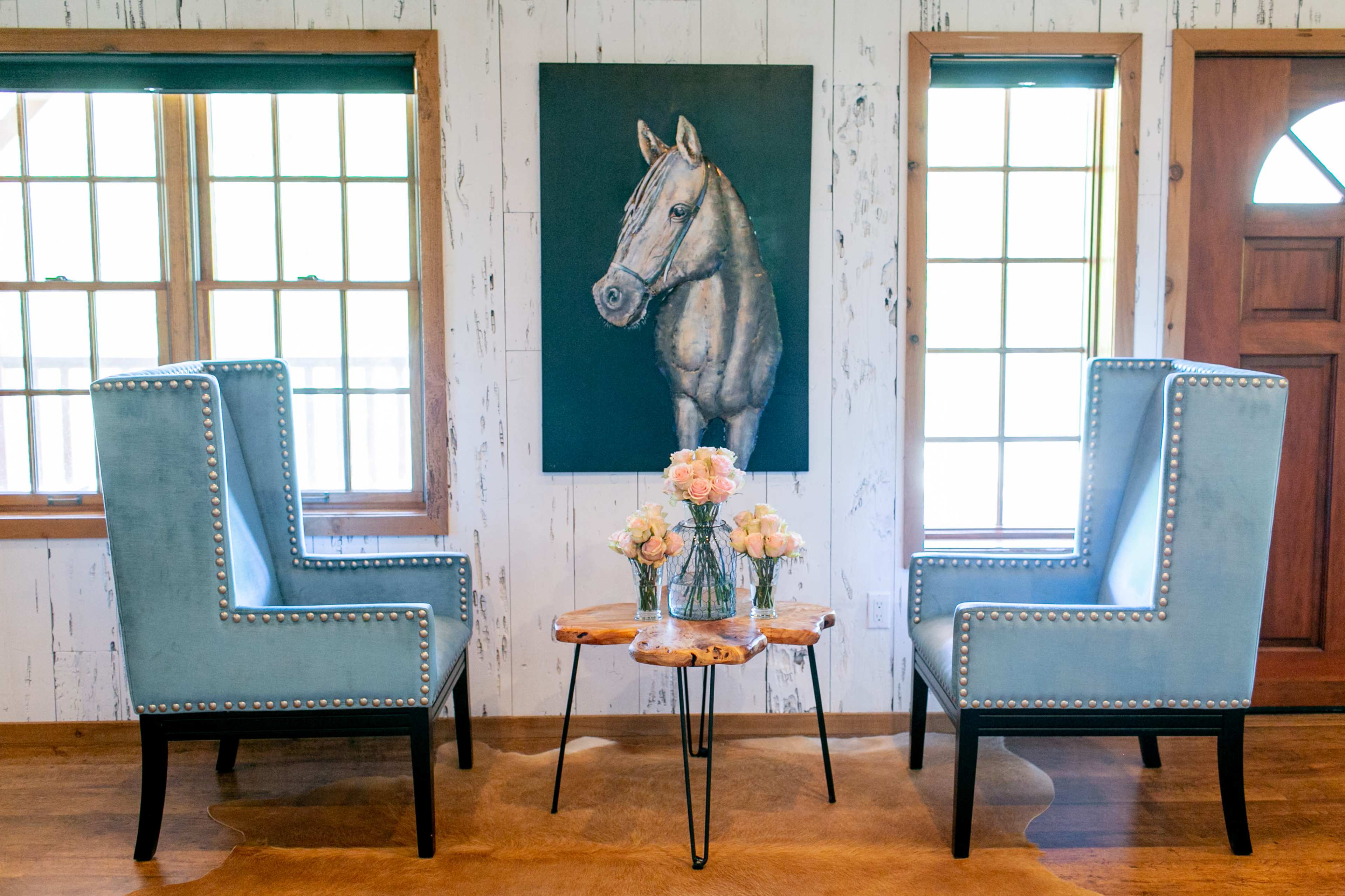 The image shows a cozy sitting area with two blue upholstered armchairs, a small wooden table, and a painting of a horse on the wall, flanked by windows with natural light.