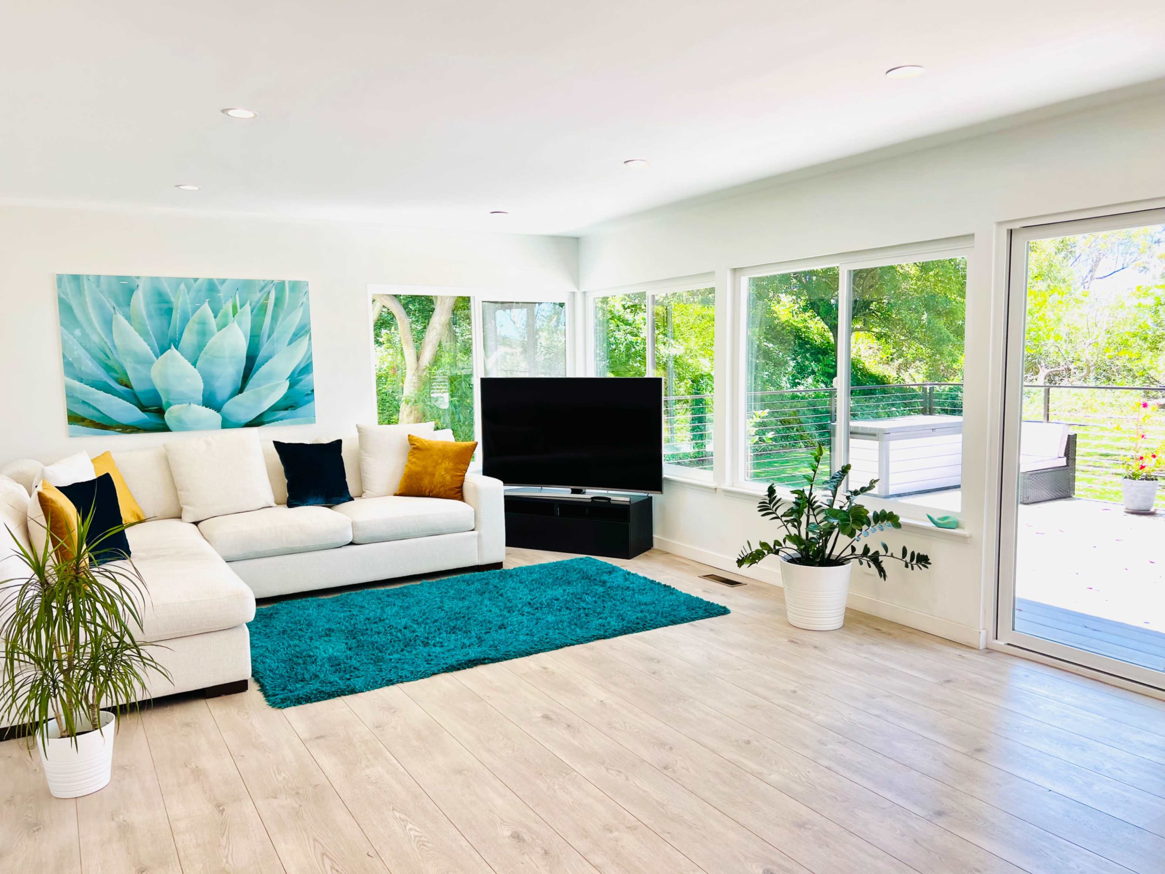 The living room features a white sectional sofa, a large black television, and a vibrant blue area rug on light wooden flooring, with large windows allowing natural light and views of greenery outside.