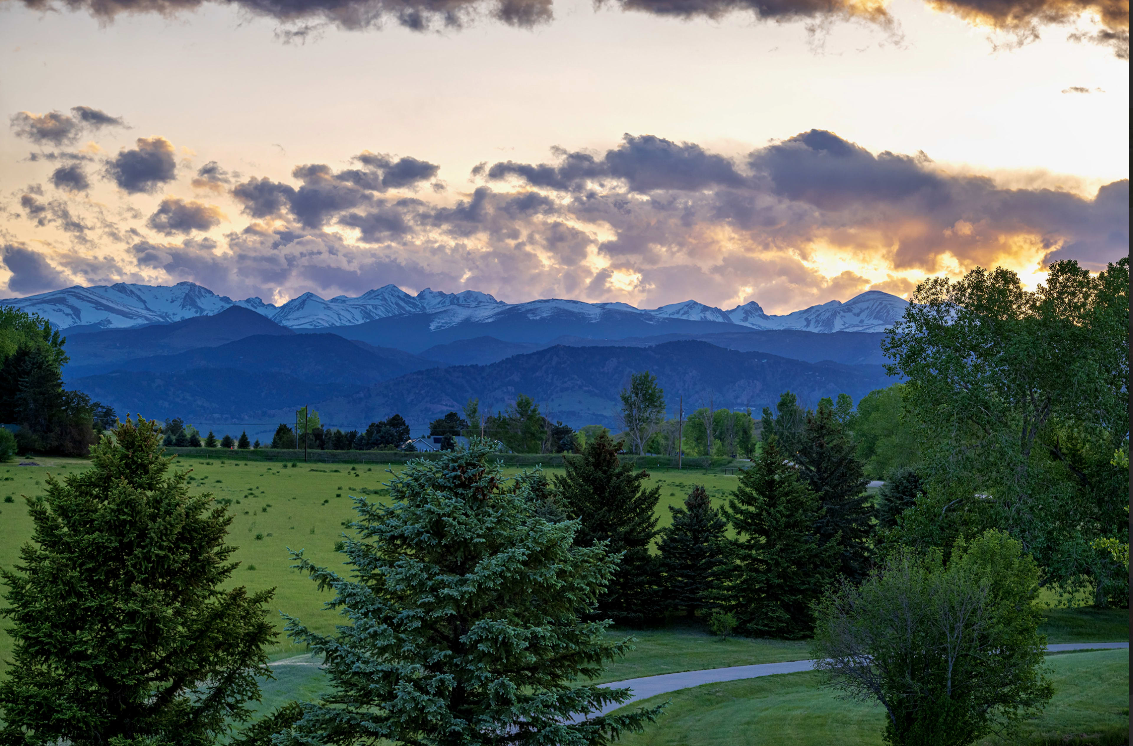 The image shows a sunset over a mountainous landscape with green fields and trees in the foreground.