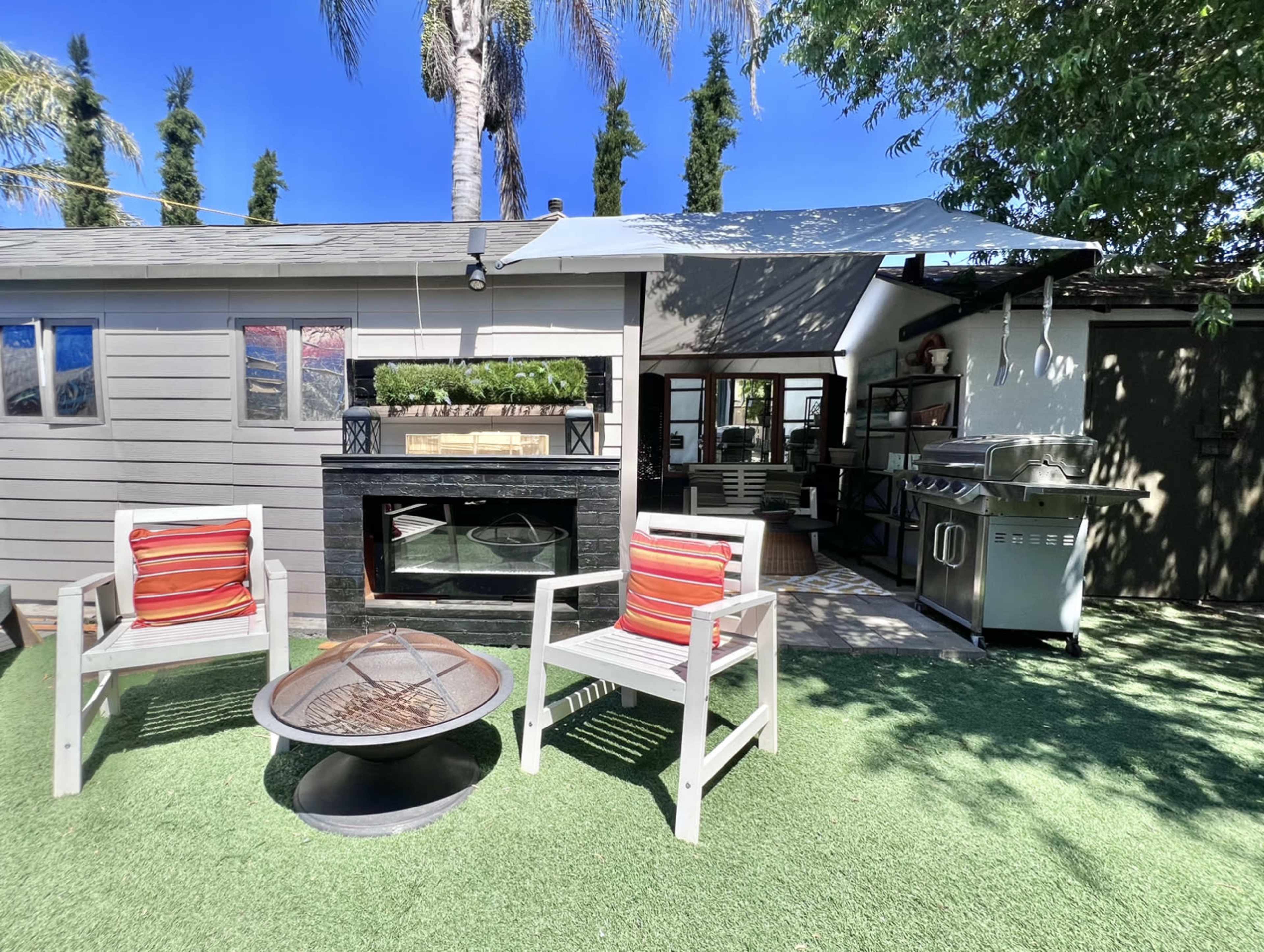 A small outdoor patio features two striped chairs, a fire pit, and a well-equipped kitchen area adjacent to a gray-shingled house.