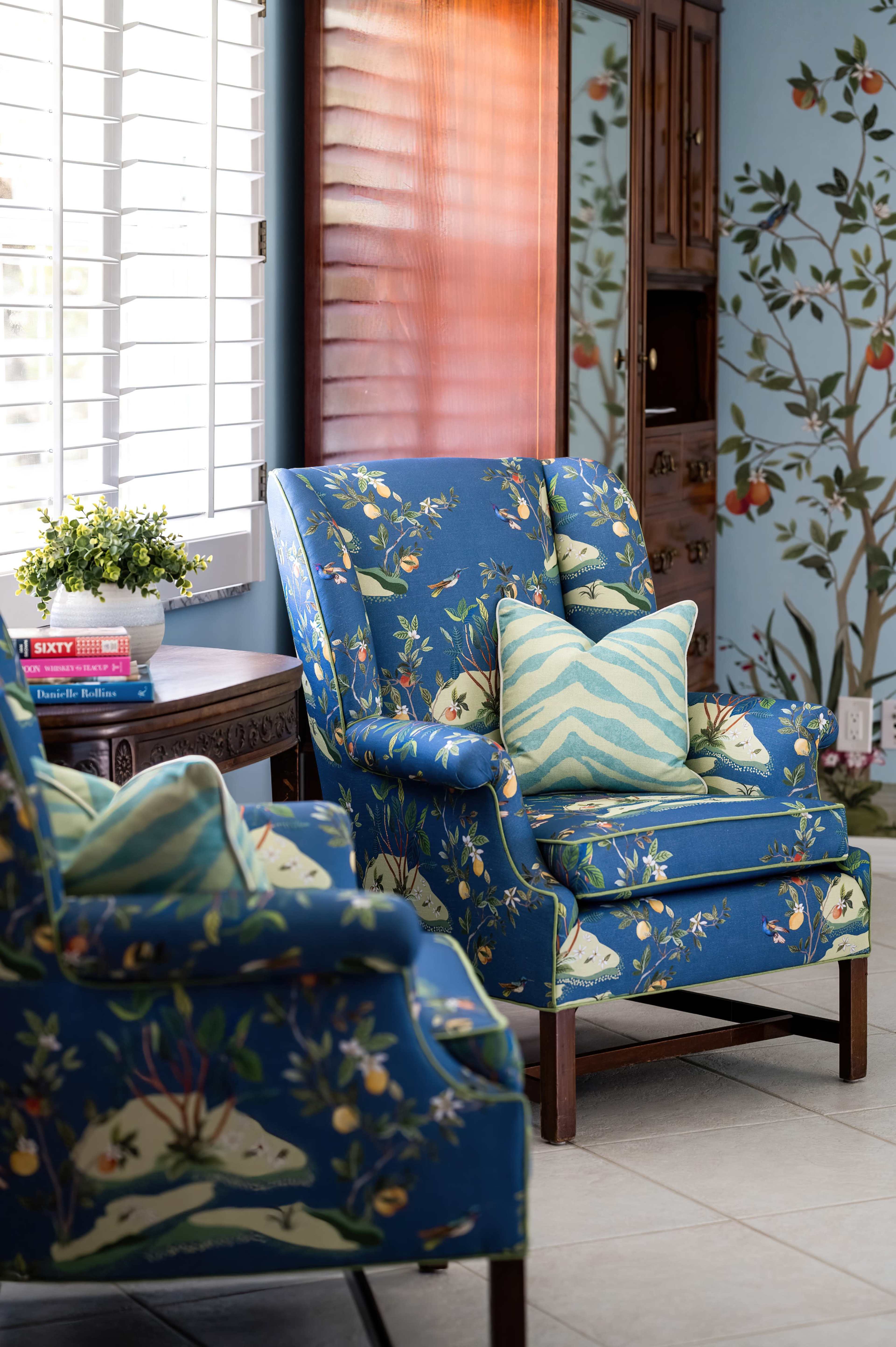 Two upholstered blue chairs with floral patterns, placed beside a wooden table in a room adorned with nature-themed wallpaper and a window allowing natural light.
