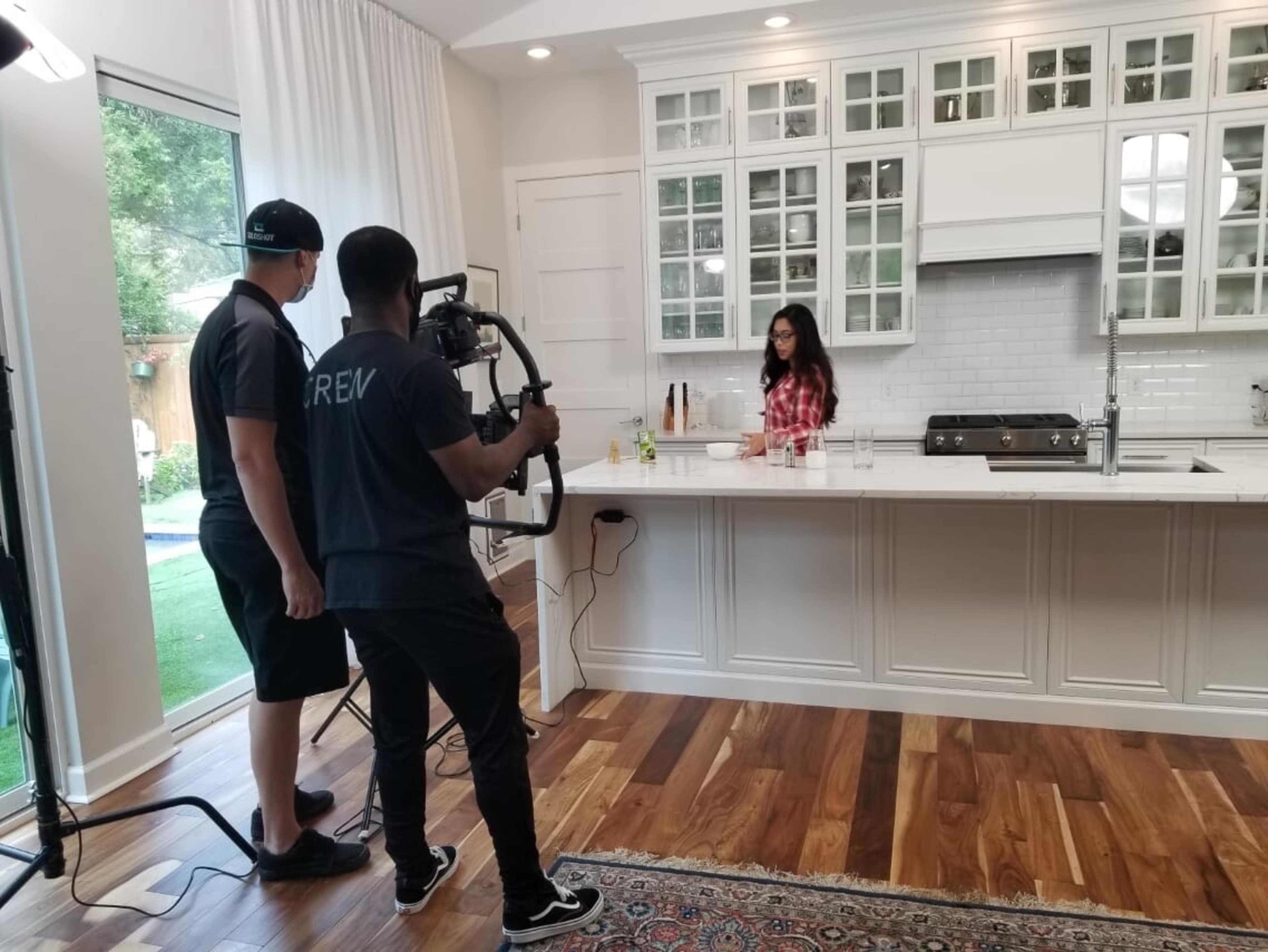 A woman prepares food at a kitchen island while two crew members film the scene with a camera setup.