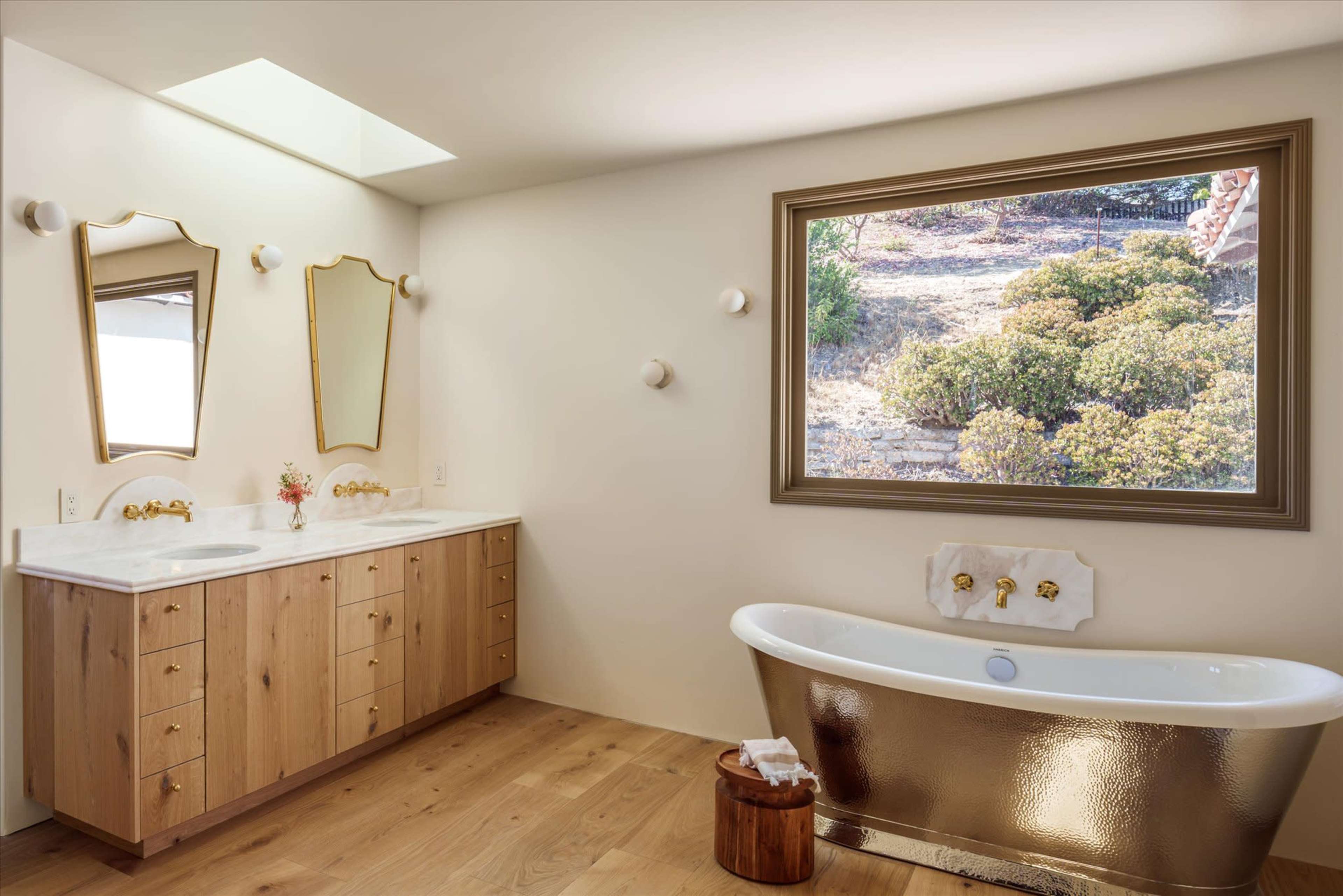 The bathroom features dual sinks with a wooden vanity, a freestanding bathtub, and a large window overlooking a natural landscape.