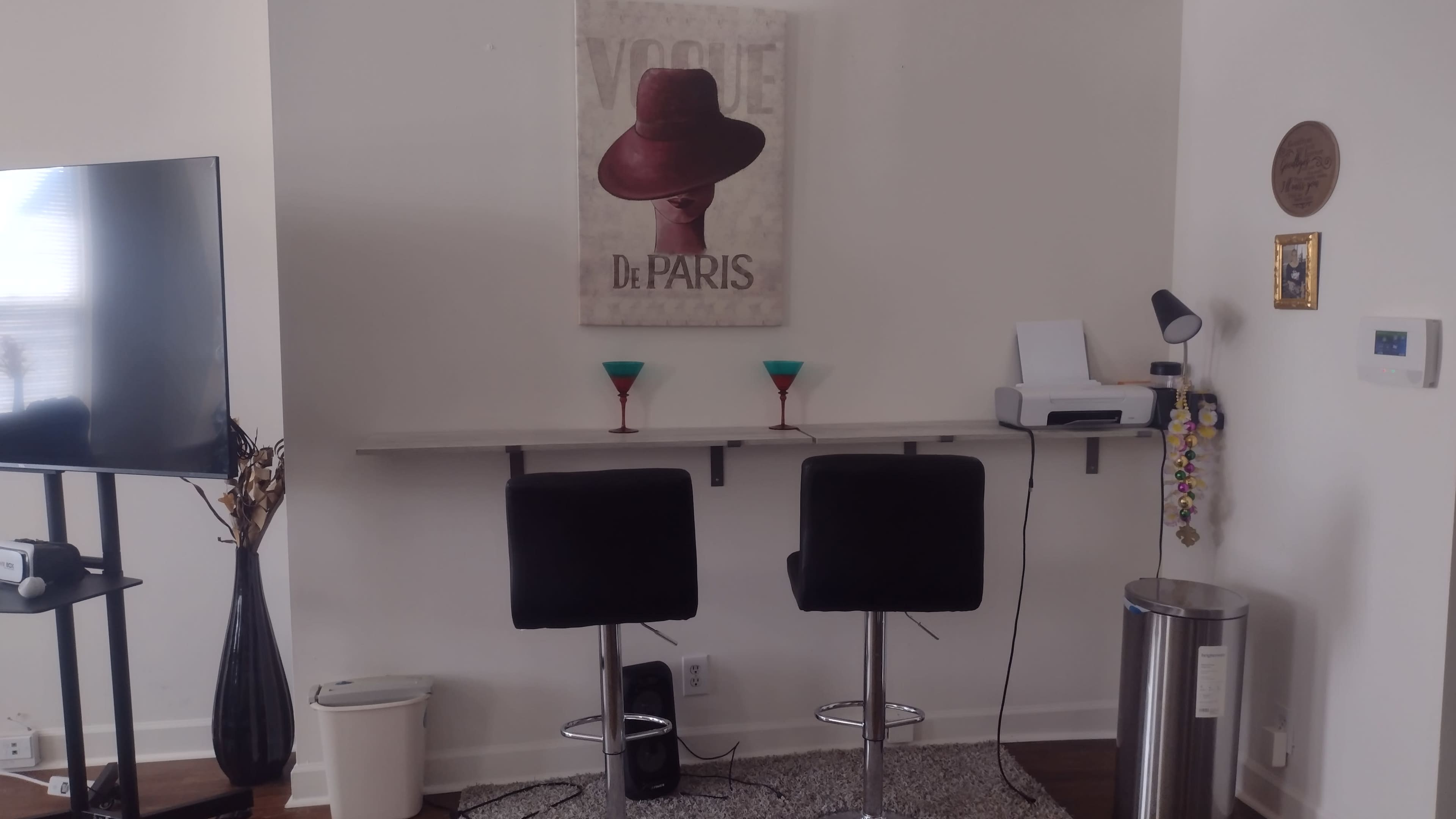 The image shows a minimalist bar area with two black stools, a wall-mounted artwork of a hat, and two colorful cocktail glasses on the counter.