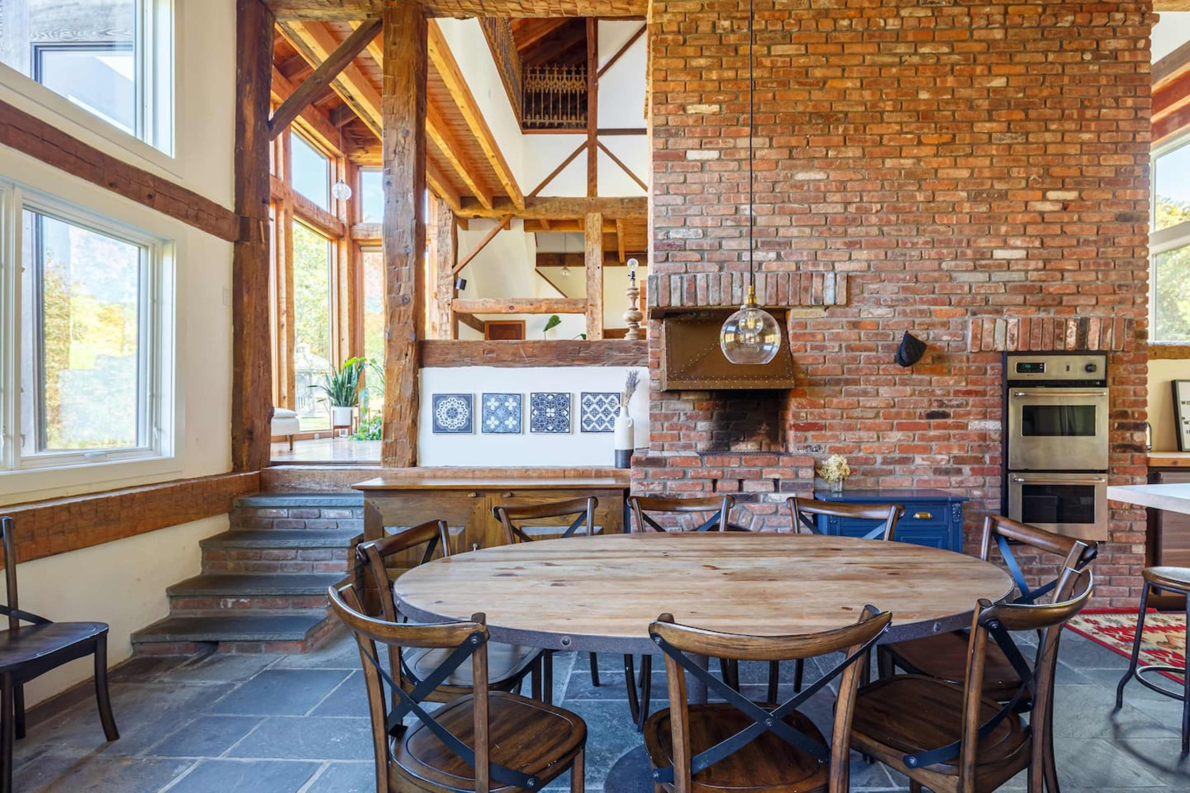 A rustic kitchen with a round wooden table surrounded by dark chairs, featuring exposed brick walls and wooden beams.