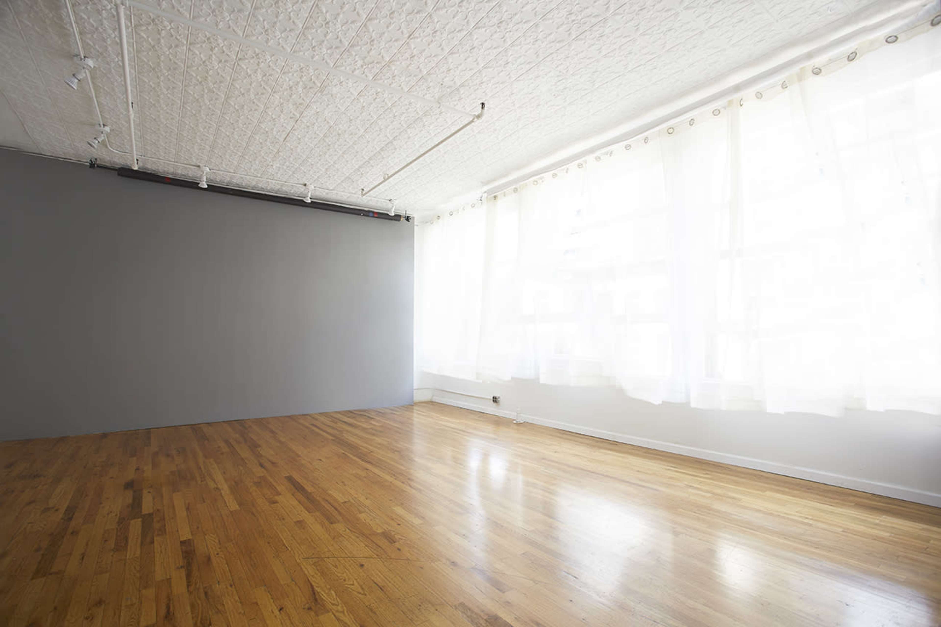 The image shows a spacious room with wooden flooring, a gray accent wall, and large windows covered with sheer white curtains.