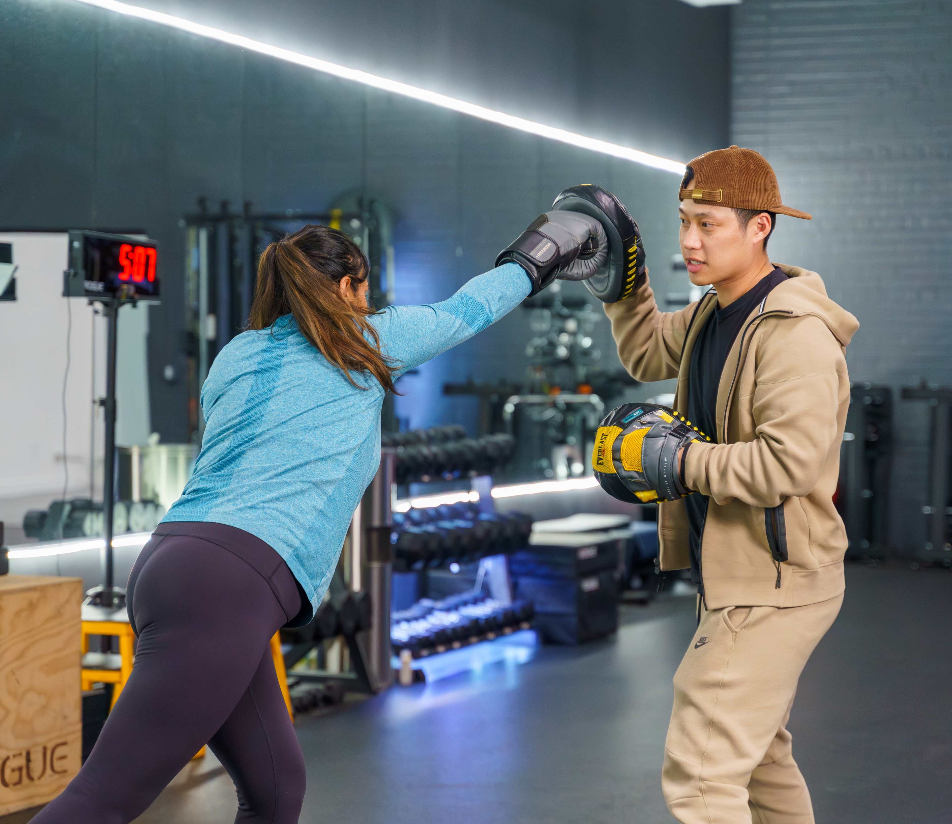 A woman in athletic wear throws a punch while practicing boxing with a trainer who is holding focus mitts in a gym.