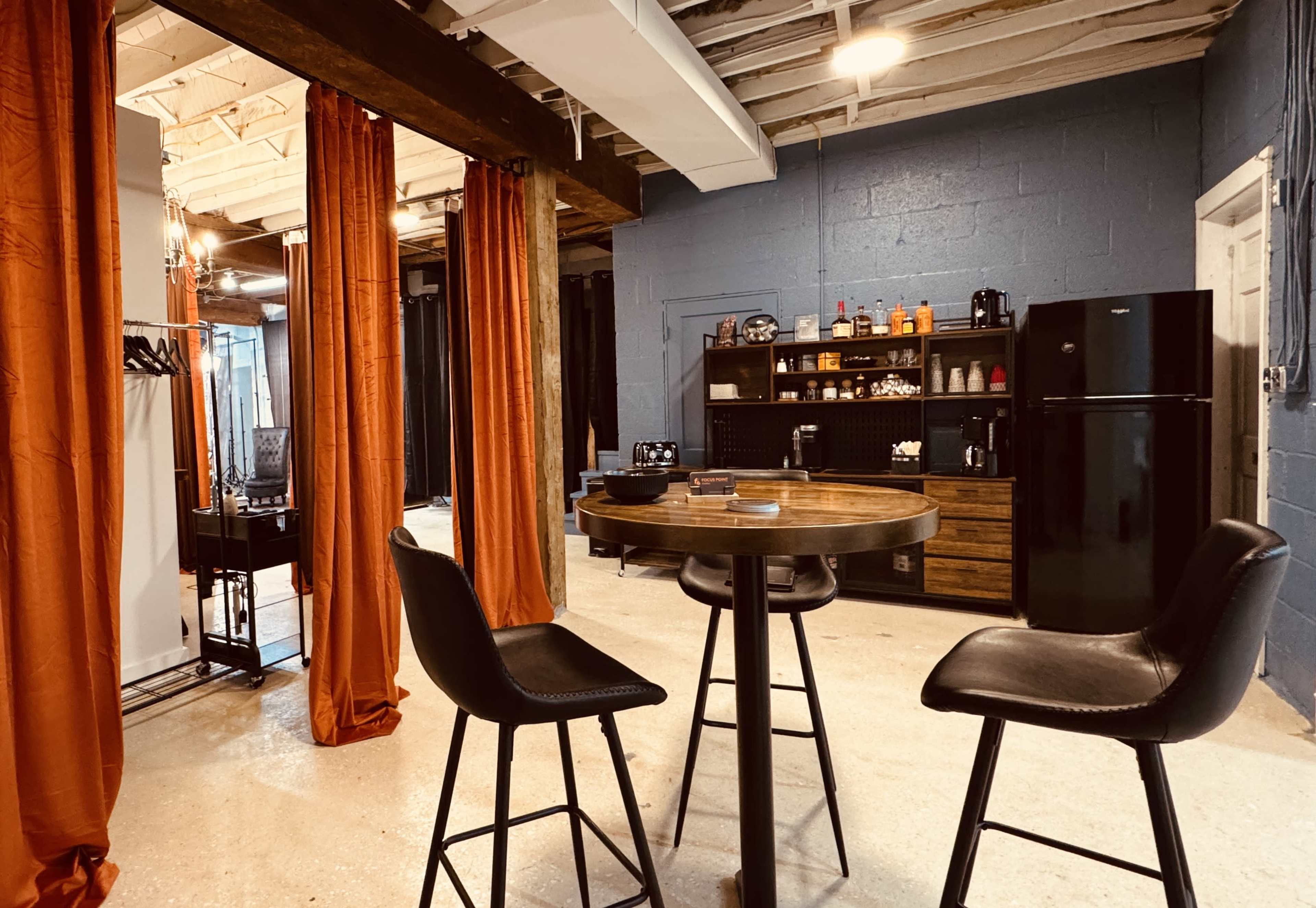 The image shows a modern, minimalist space with a circular table and two black stools, flanked by orange curtains and featuring a kitchen area with a black refrigerator and shelves displaying various items.