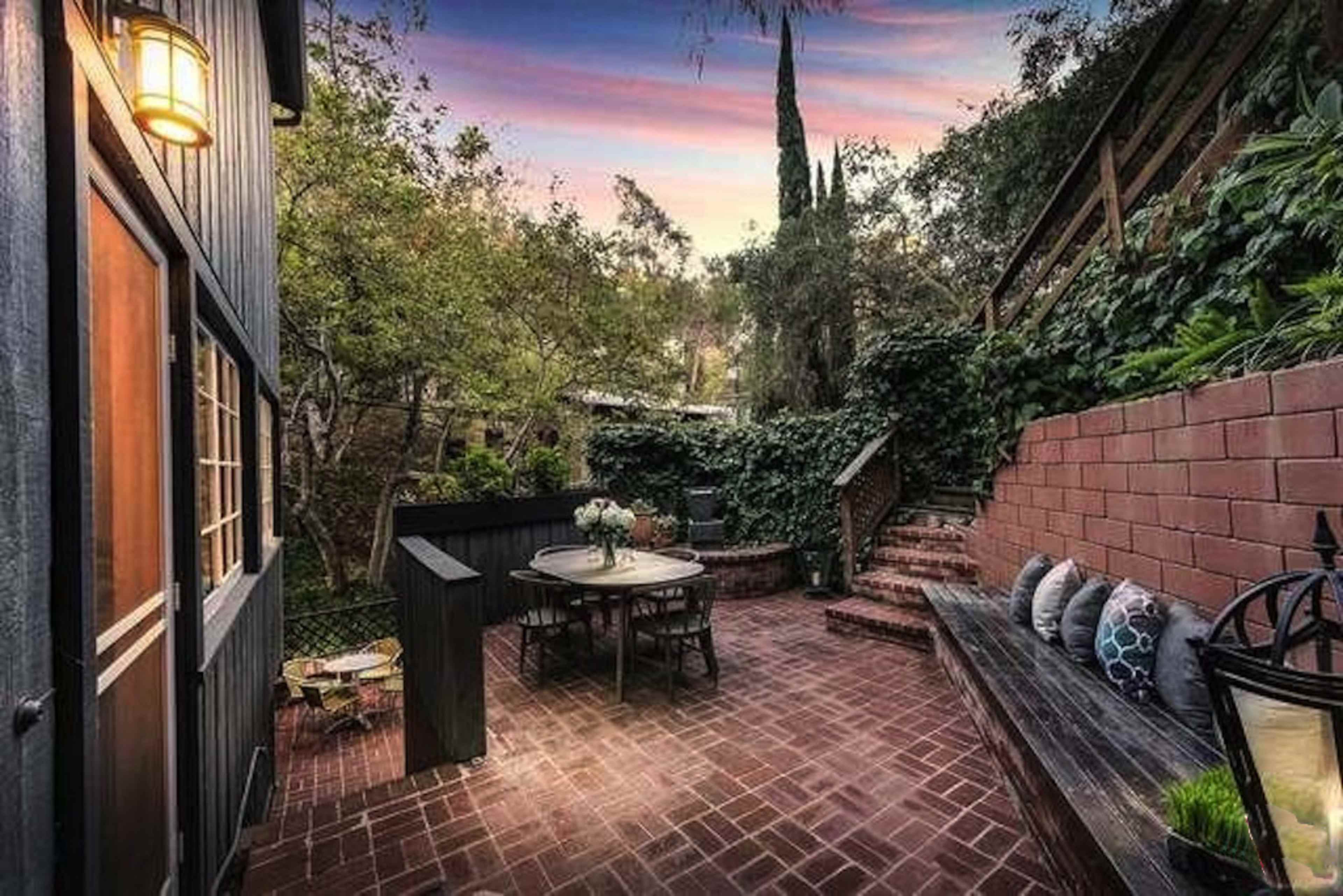 A brick patio with a table and chairs is surrounded by greenery and walls covered in climbing plants, under a colorful sky at dusk.