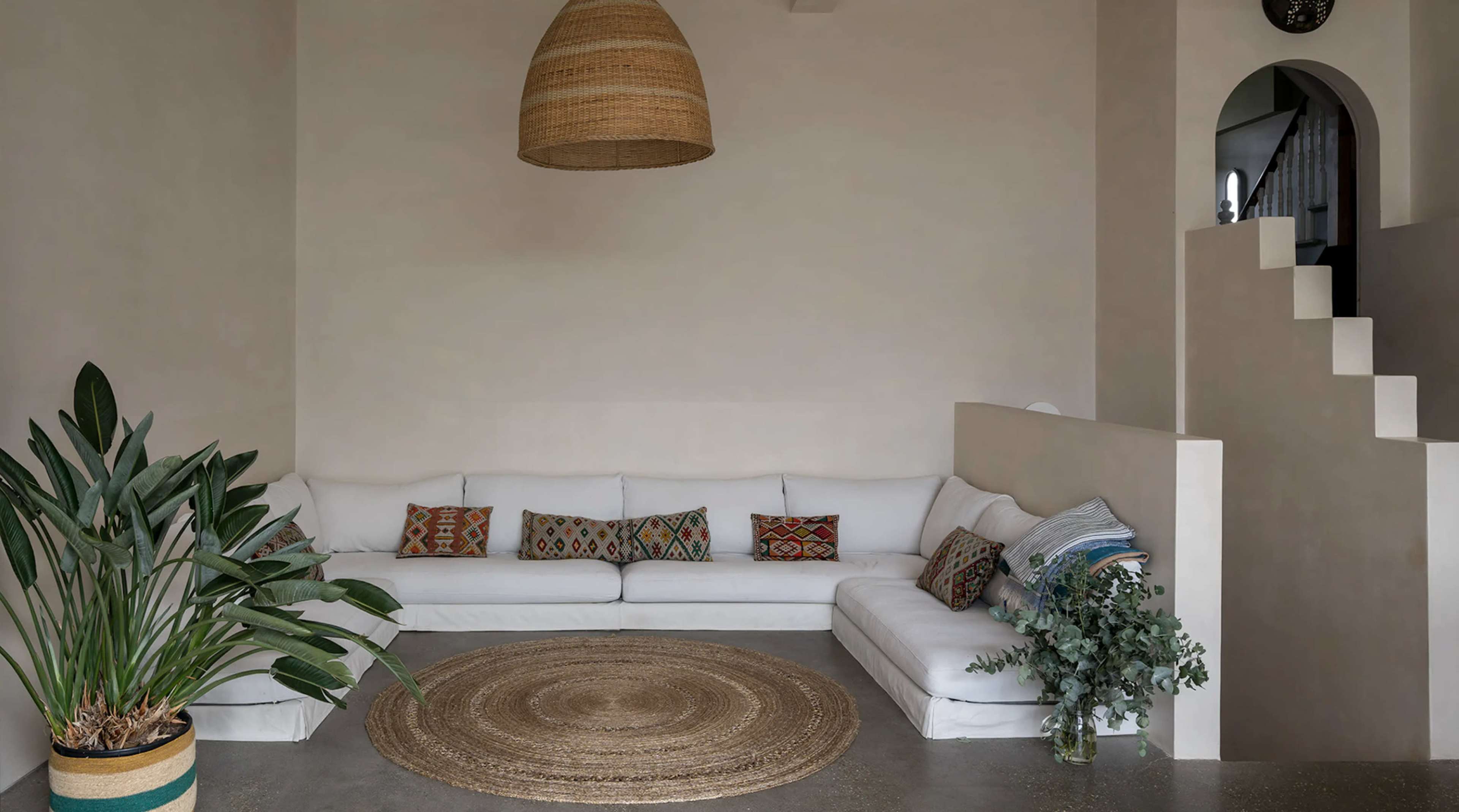 A minimalist living room featuring a white sectional sofa, a round jute rug, and a large plant, with a staircase in the background.