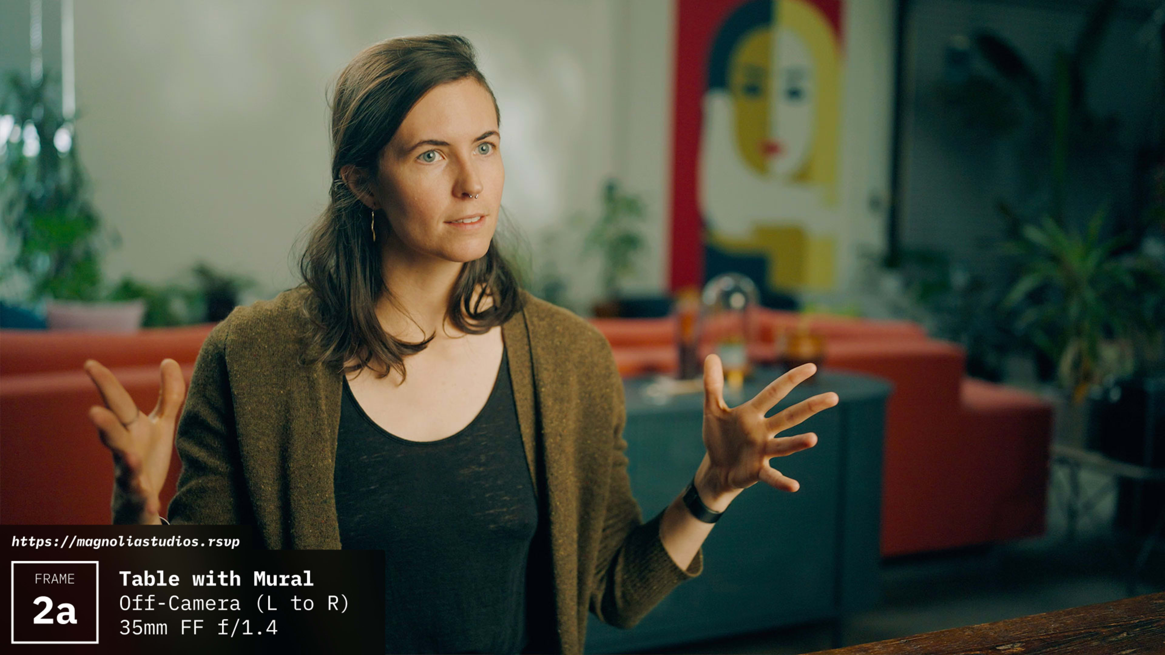 A woman is gesturing with her hands while speaking, seated at a table in front of a colorful mural.