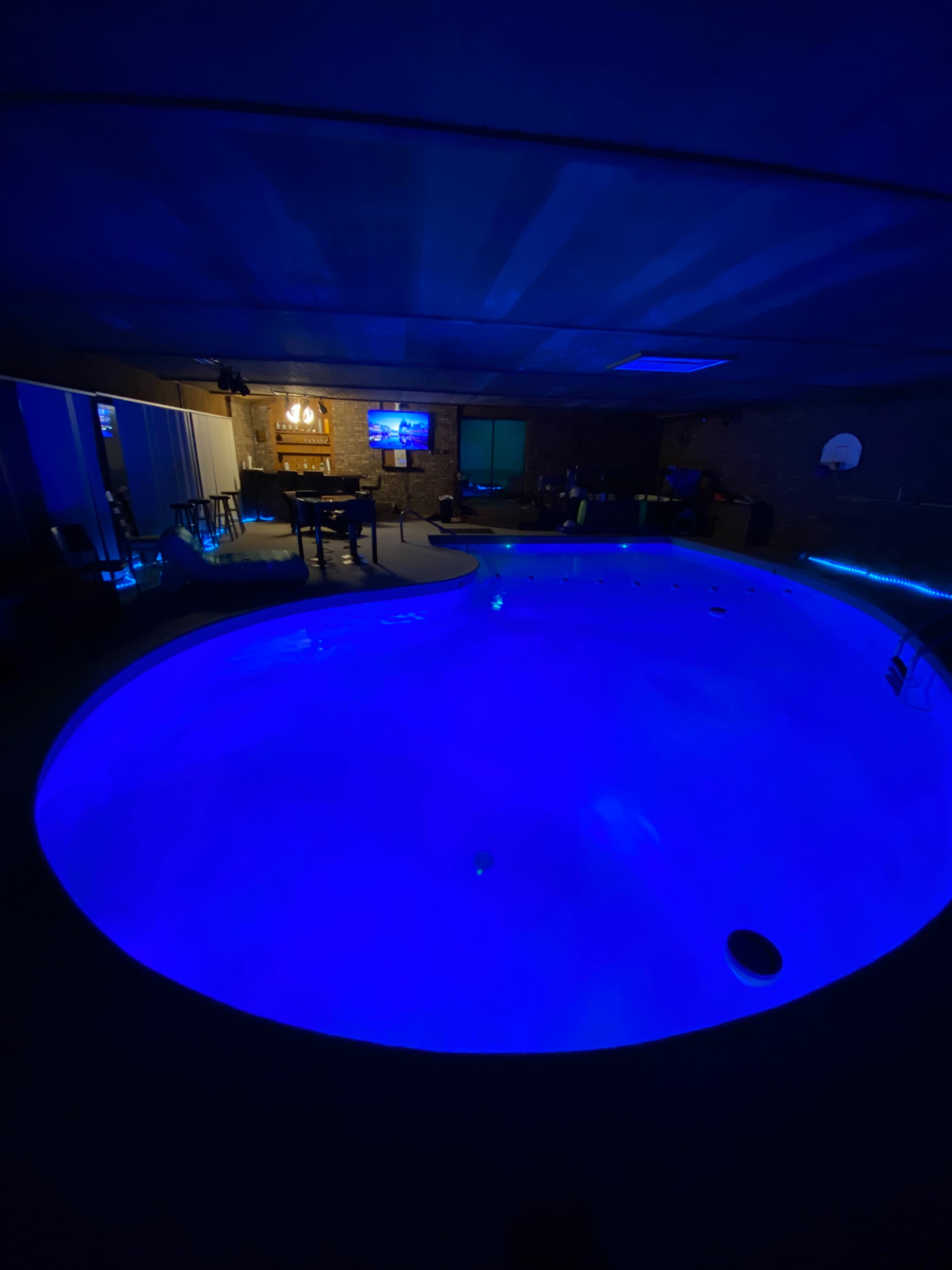 An indoor pool illuminated with blue lighting, surrounded by a dimly lit room featuring a bar area and seating.