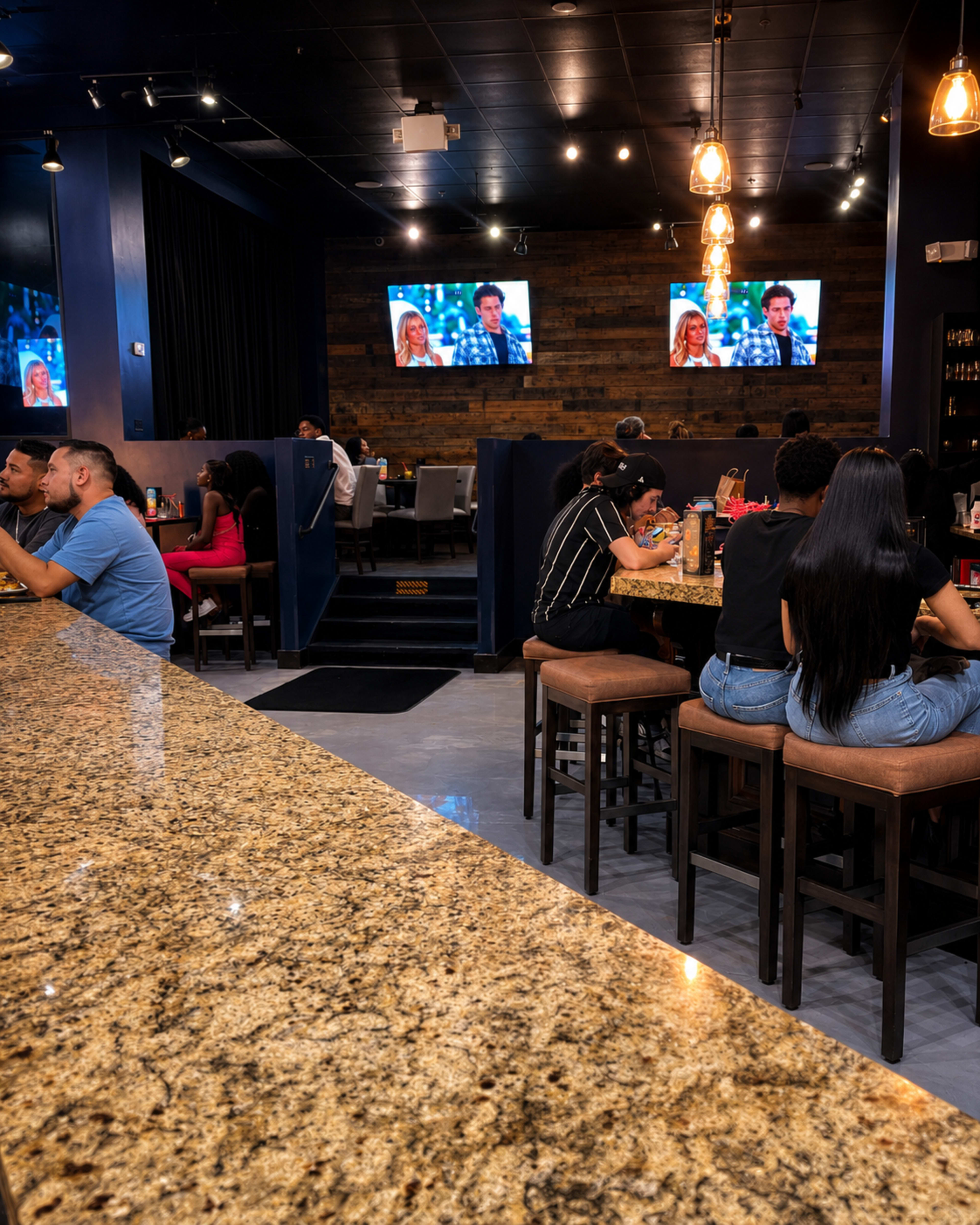 The image shows a restaurant interior with patrons seated at the bar and tables, and two television screens displaying images on the walls.