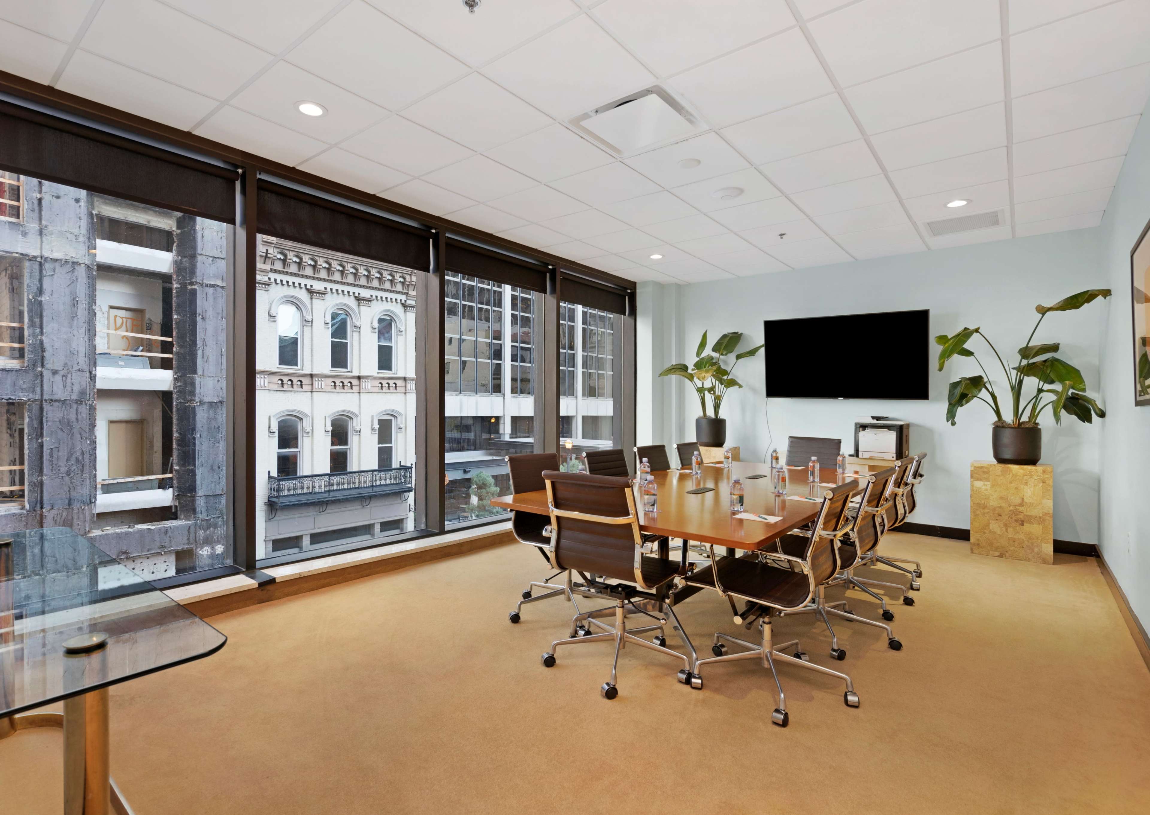 A conference room features a long table surrounded by chairs, with large windows offering a view of buildings outside, and a television mounted on the wall.