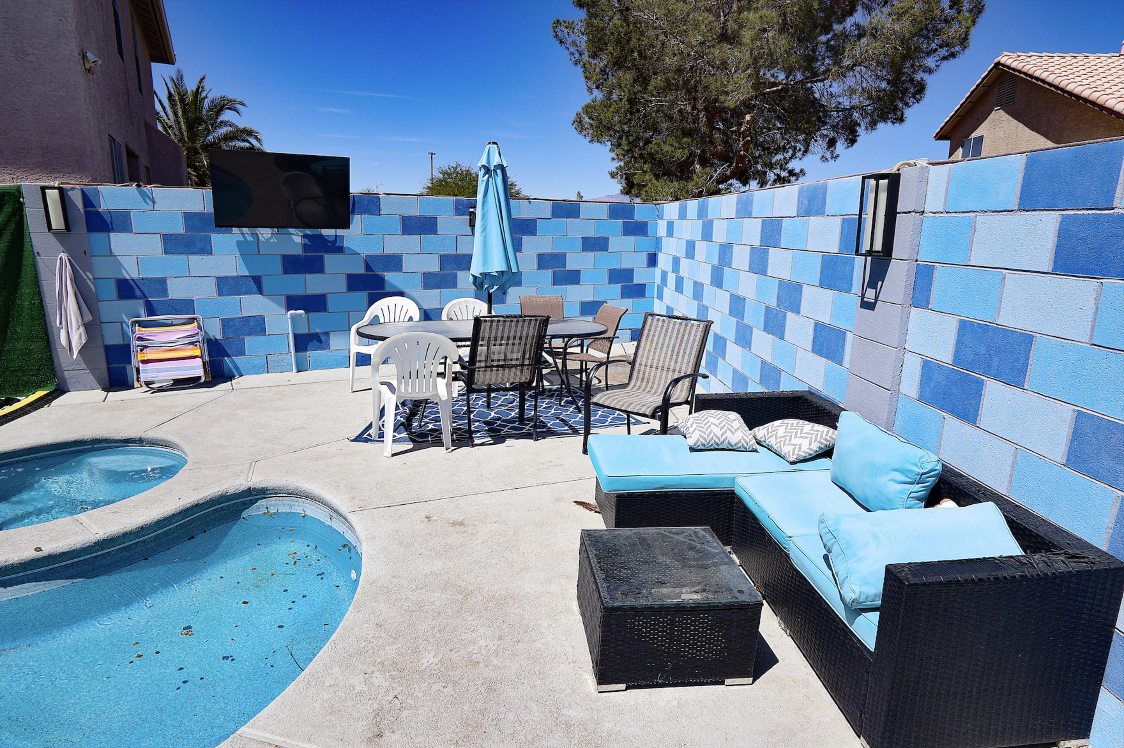 The image shows a small outdoor patio area with a swimming pool, seating furniture, a dining table, and a blue umbrella surrounded by a colorful block wall.