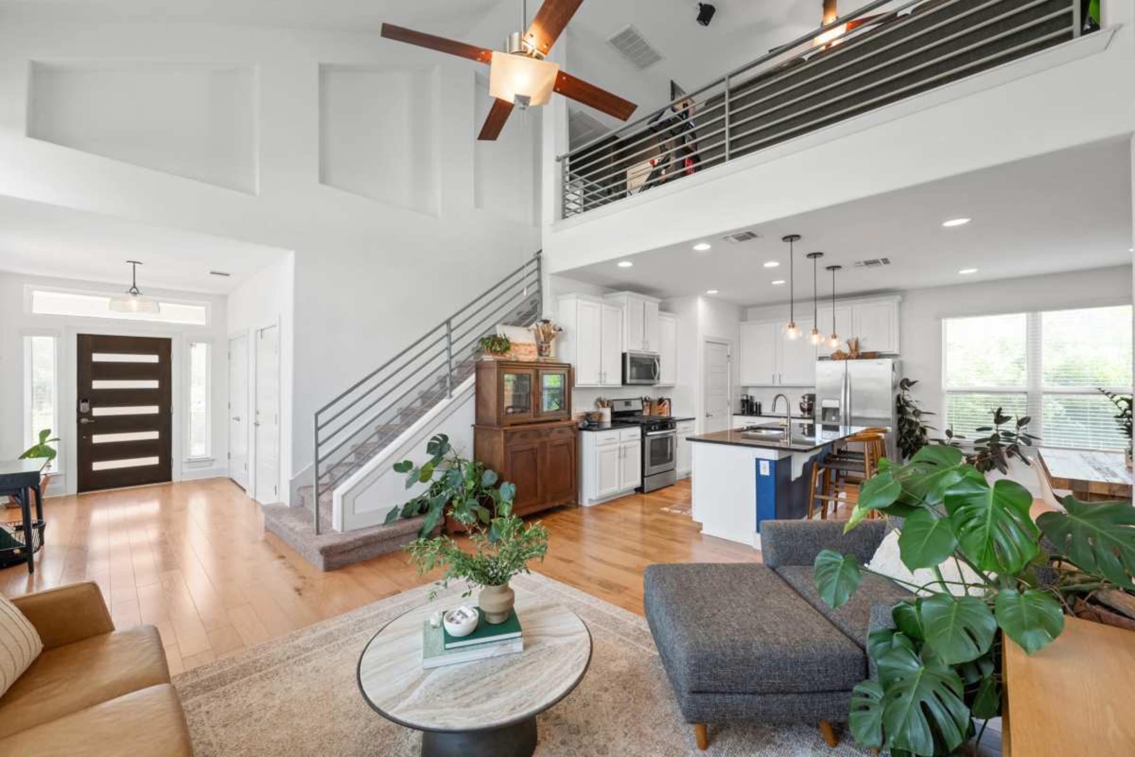 The image shows a bright, open-concept living area featuring a sofa, a coffee table, and a staircase leading to an upper level with a kitchen visible in the background.