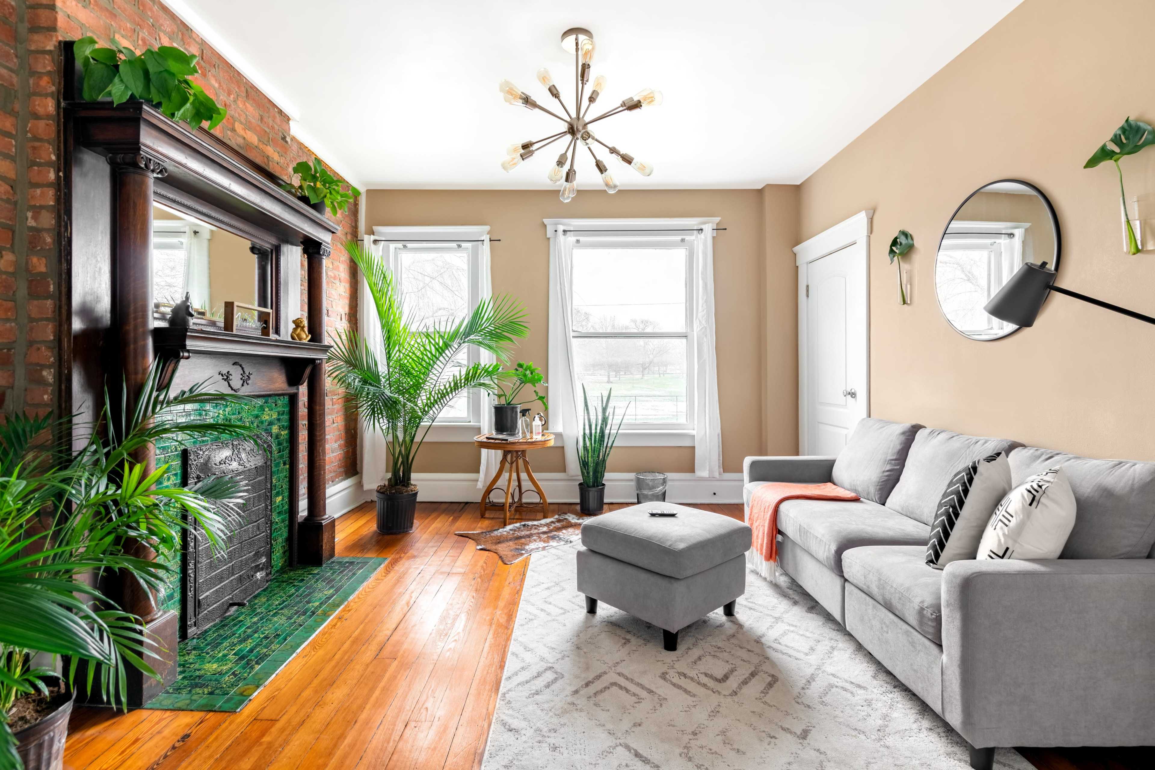 A living room with a gray sofa, a decorative ottoman, green plants, a brick wall with a fireplace, and large windows allowing natural light.