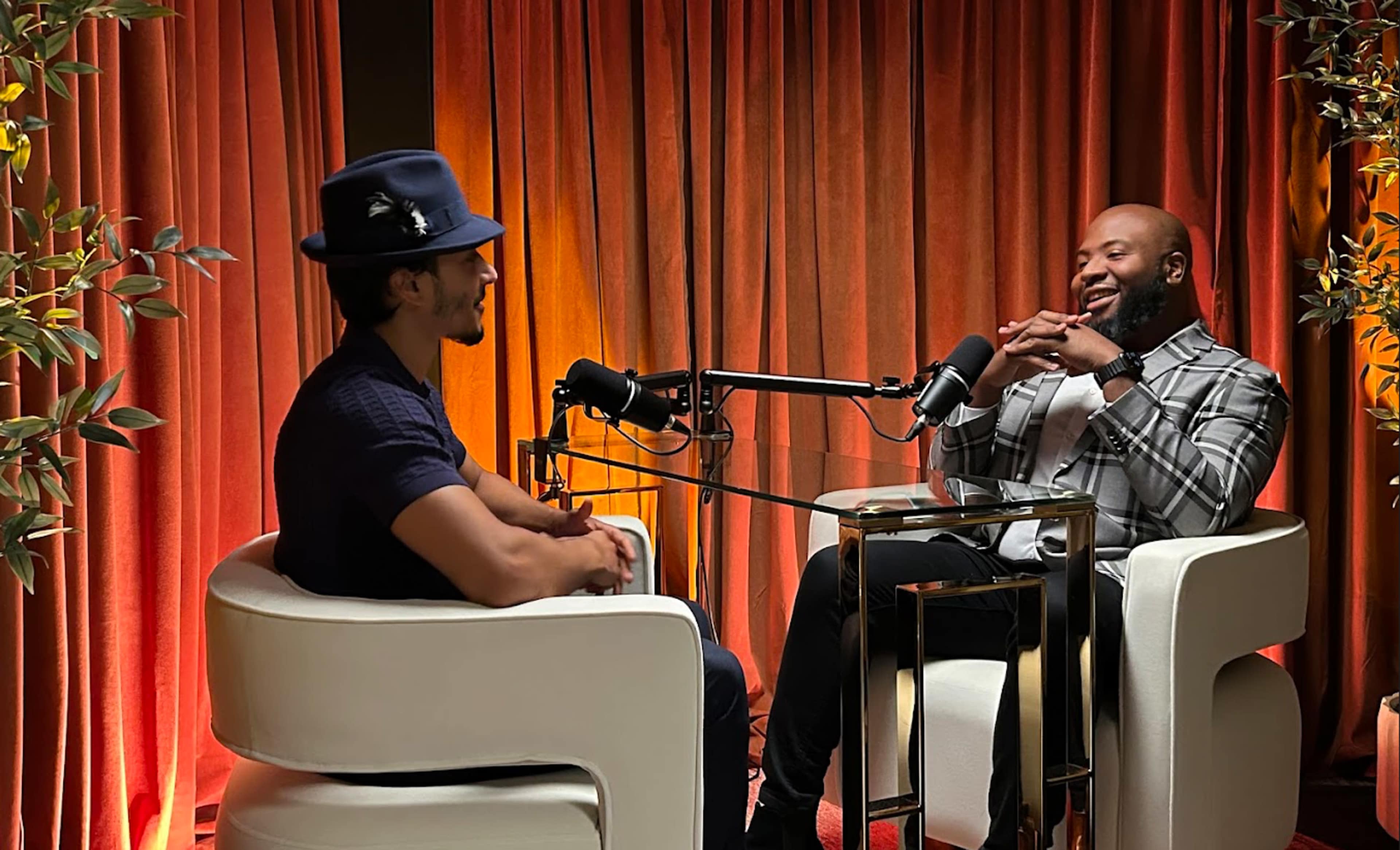 Two men are seated in modern white chairs at a glass table, engaged in conversation with microphones set up in front of them, against a backdrop of orange drapes.