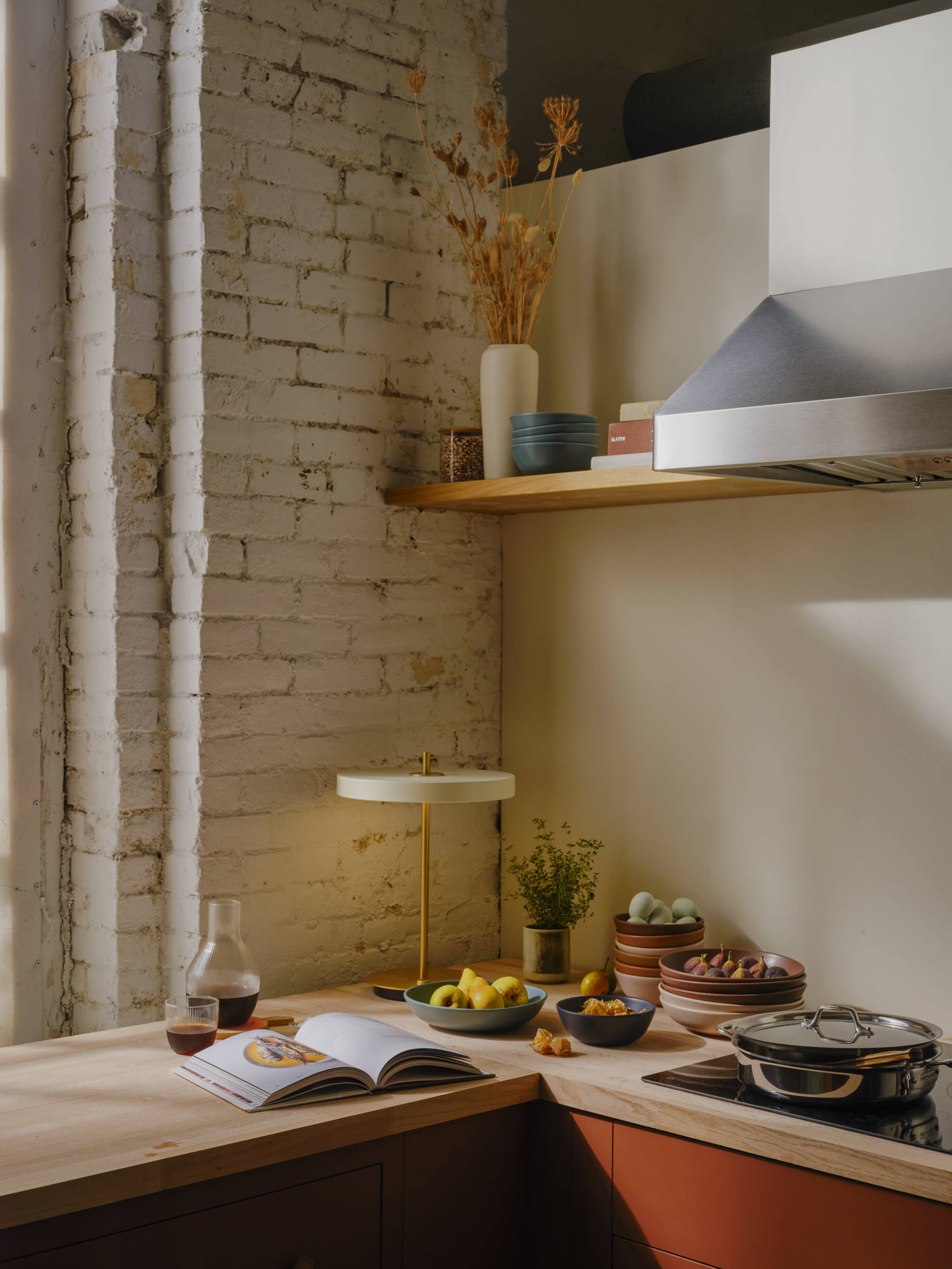 A modern kitchen corner features a wooden countertop with a cookbook open, bowls of fruit, a glass of drink, and a lamp, accompanied by decorative plants and a stainless steel hood overhead.
