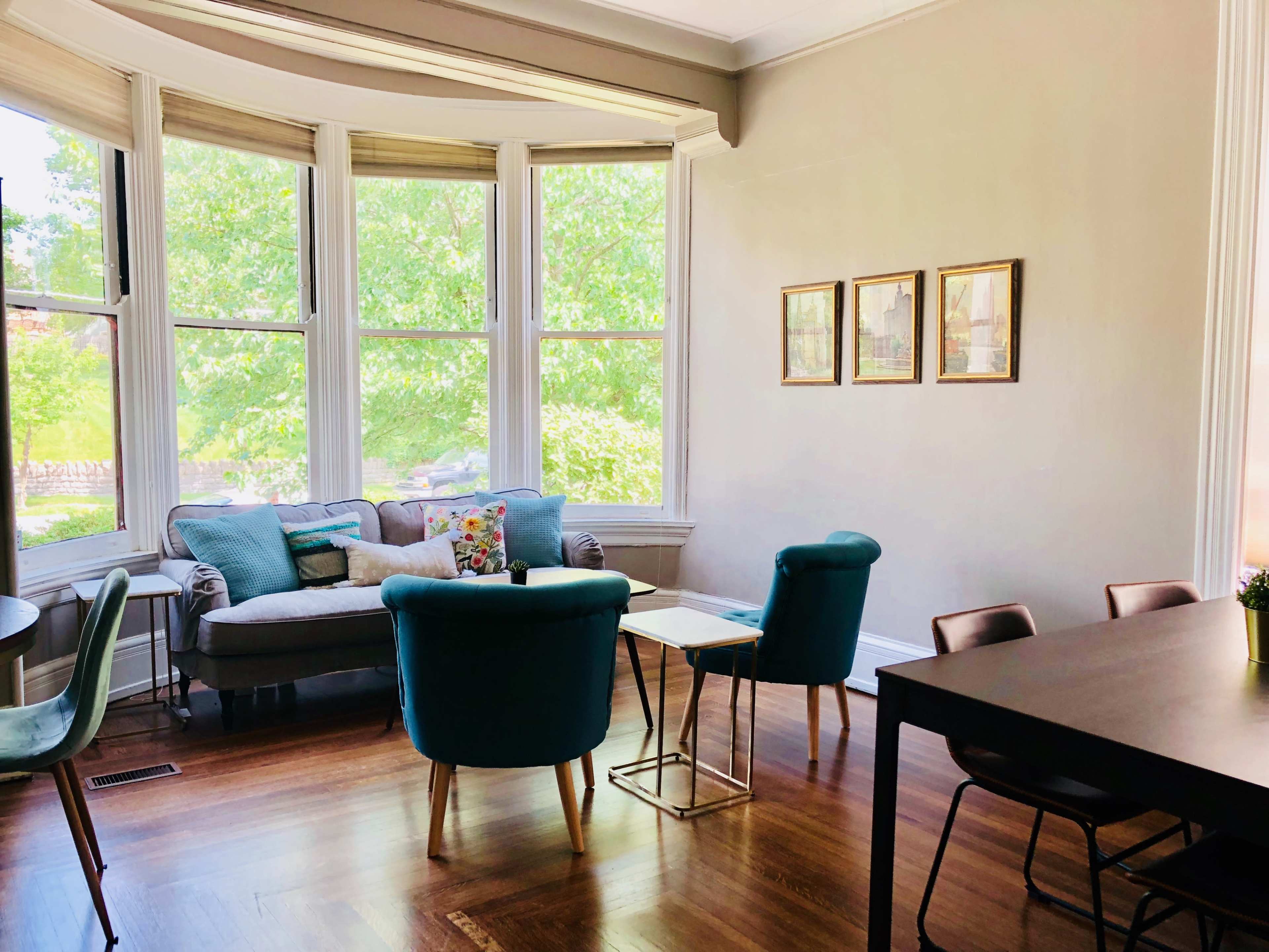 A bright, open living area features a bay window, a sofa, two teal chairs, and a dining table.