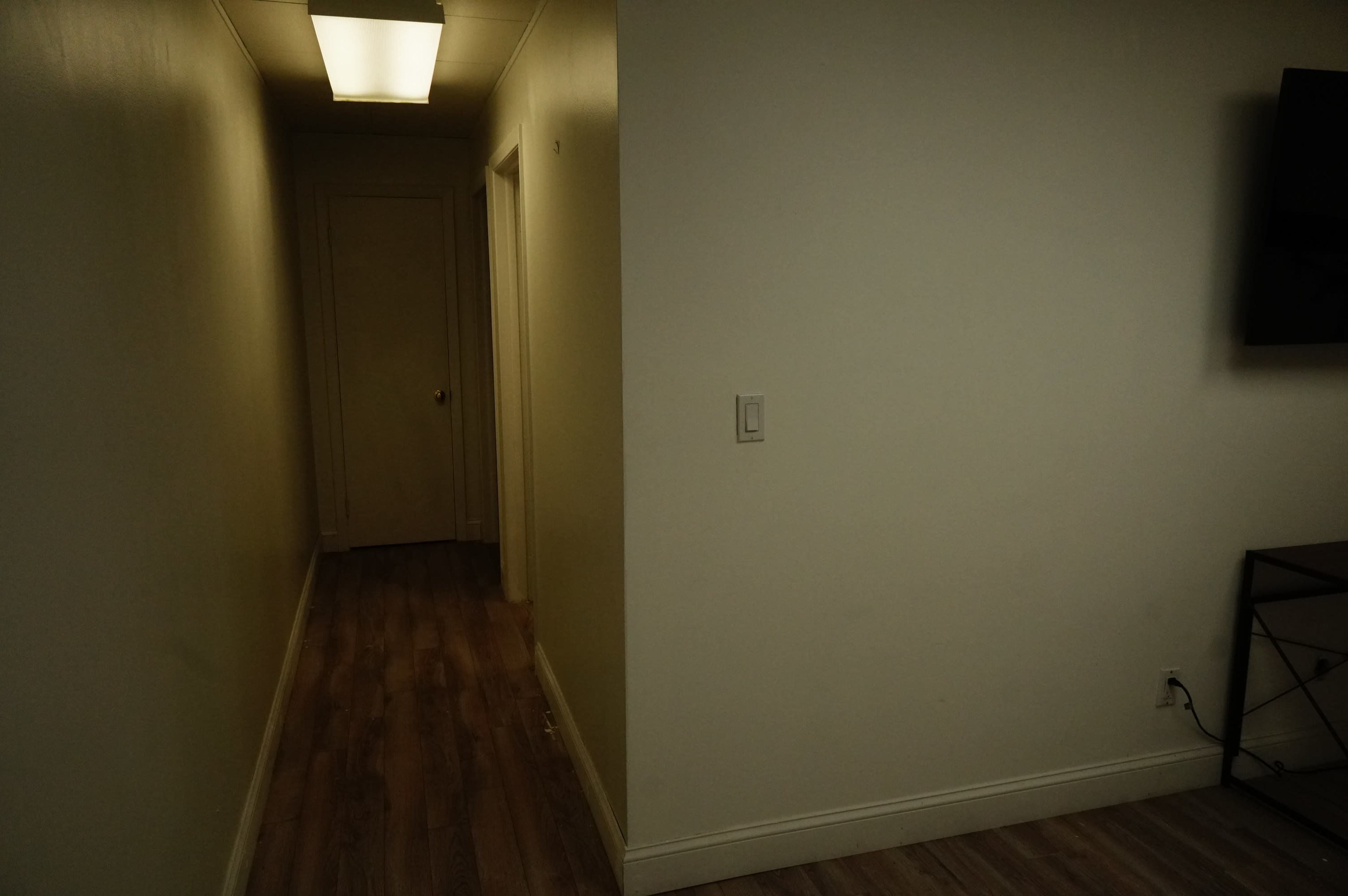 A narrow corridor leads to a closed door, with light coming from a ceiling fixture and a wall-mounted television on the right.