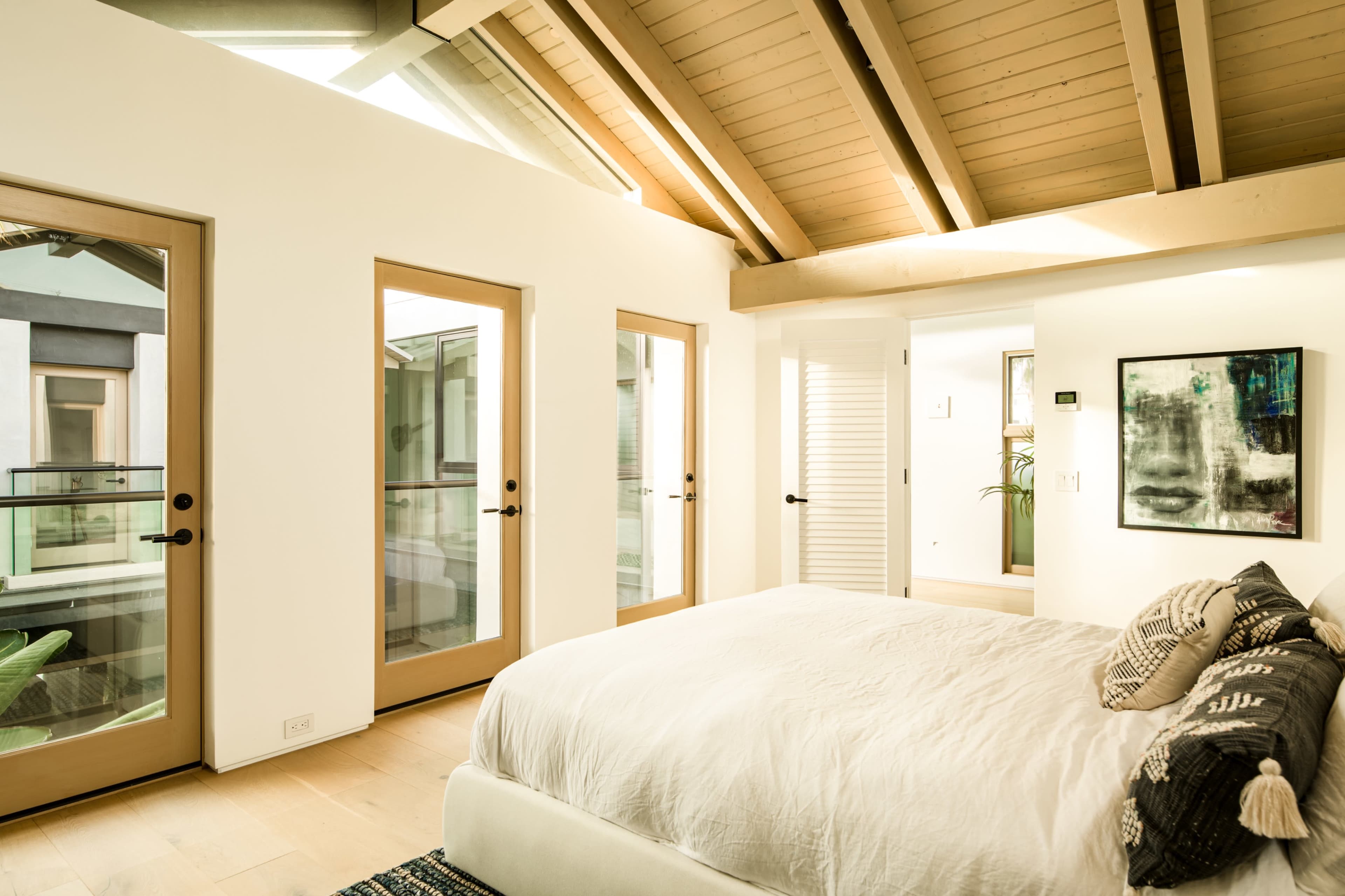 A well-lit bedroom features a large bed with a white bedding set and three doors leading to an outdoor balcony area.