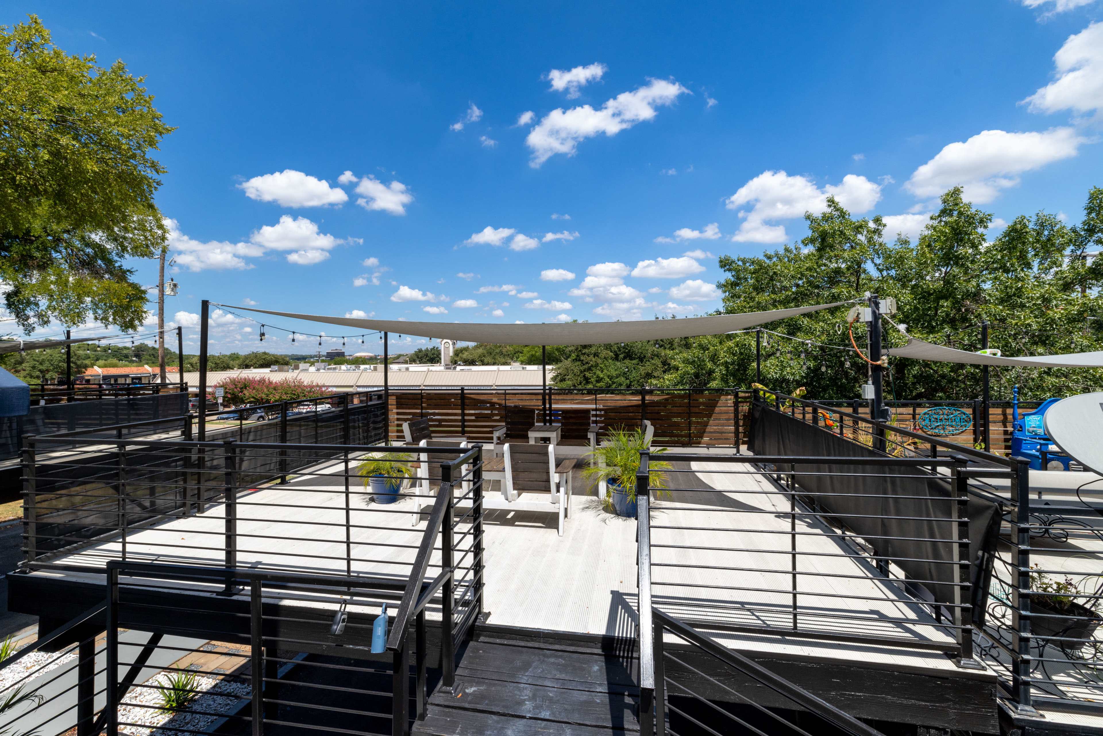 The image shows a rooftop deck with wooden flooring, lounge chairs, and shade sails, set against a clear blue sky with scattered clouds.