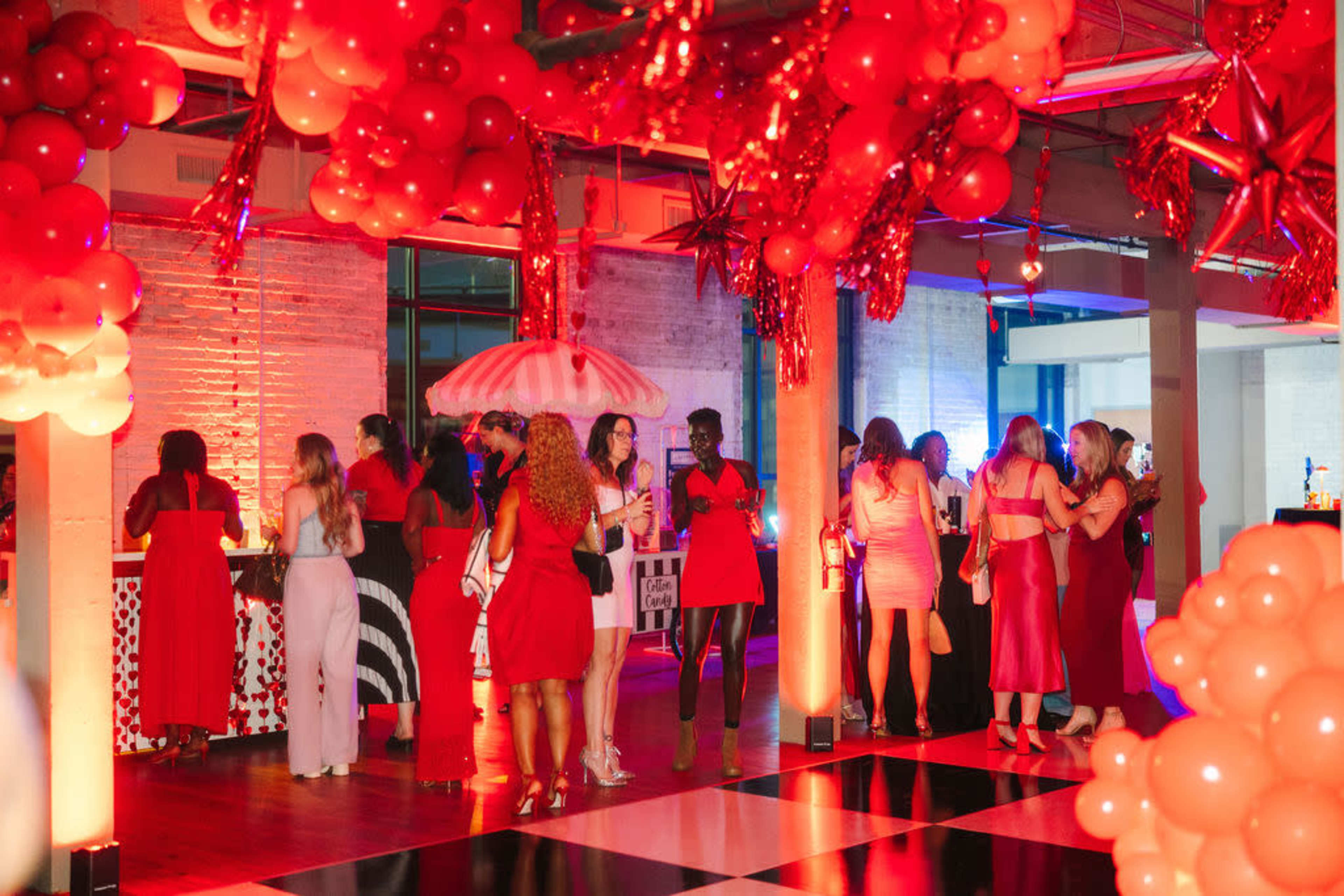 The image shows a lively event space decorated with red balloons and lighting, where attendees in various red outfits are socializing and enjoying drinks.