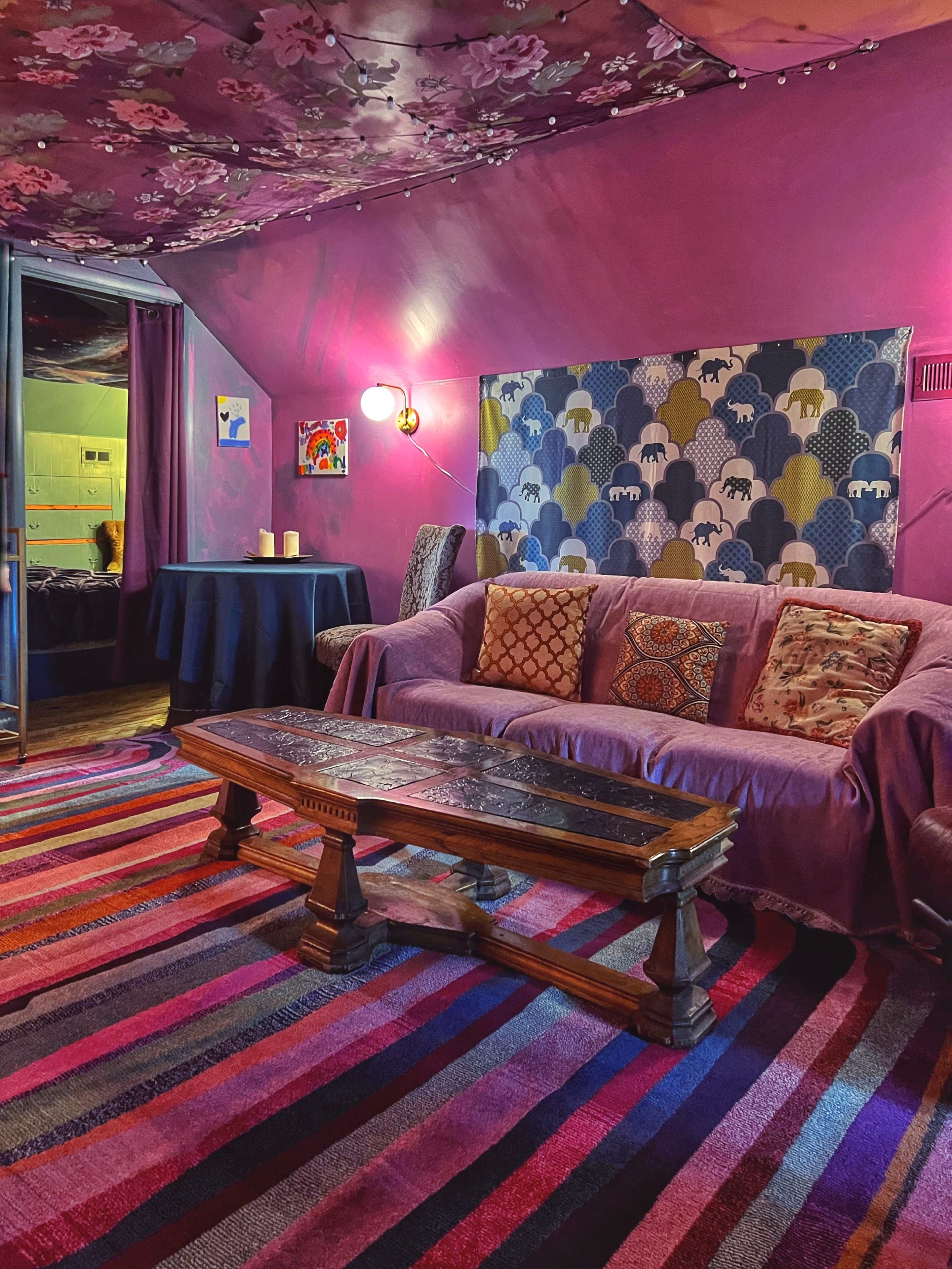 The image shows a cozy room with purple walls, a couch covered in a blanket, a wooden coffee table, and a colorful striped rug.