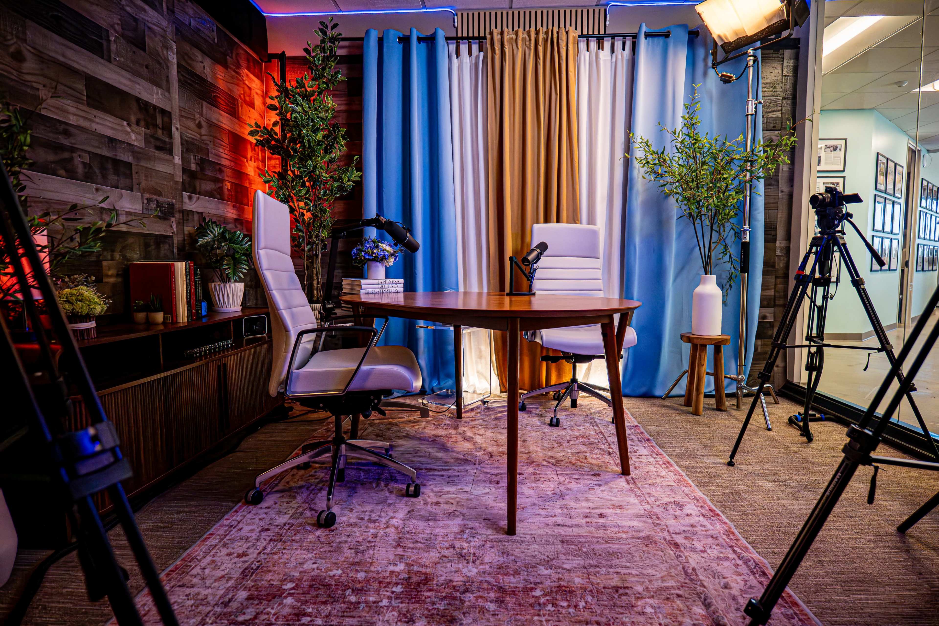 The image shows a modern studio setup with two white chairs, a wooden table, and various plants, illuminated by soft lighting and draped curtains.