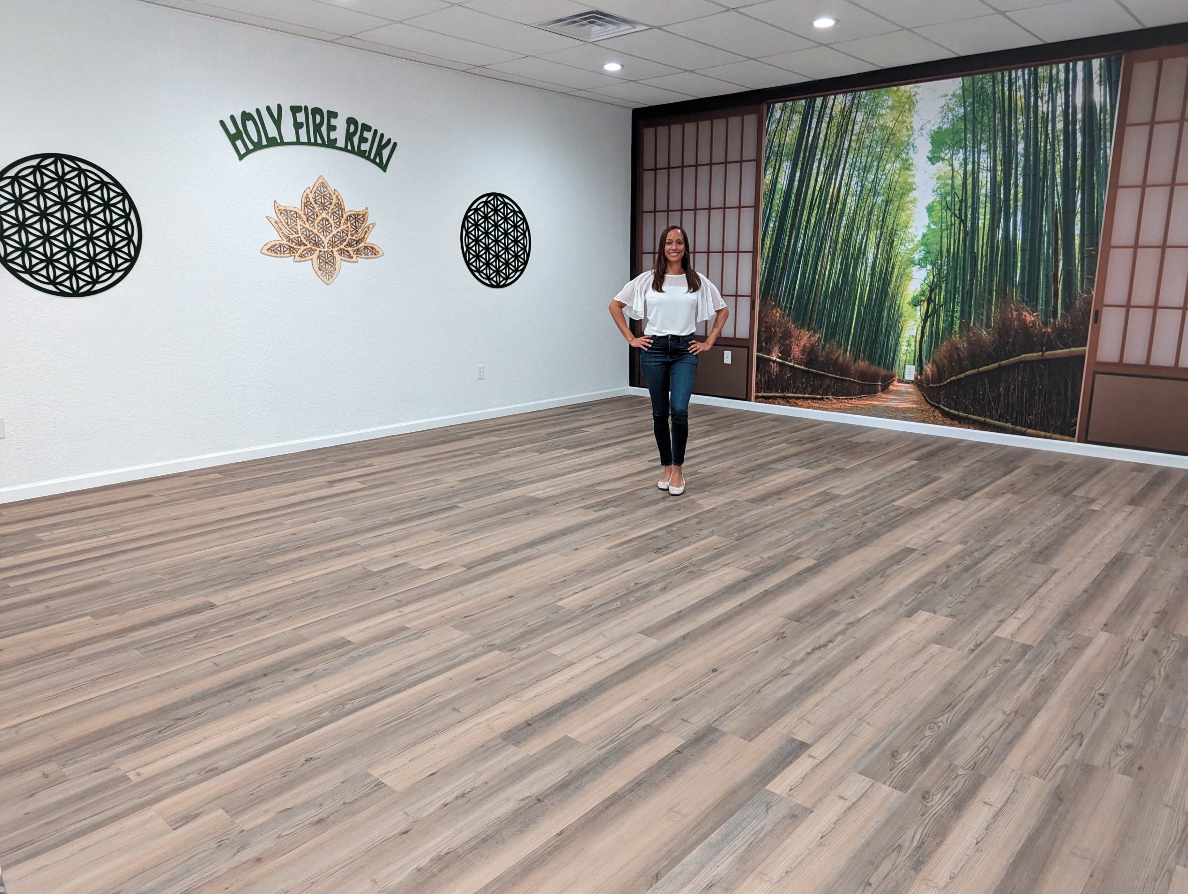 A woman stands on a wooden floor in a brightly lit yoga studio featuring a large bamboo mural and decorative wall art.